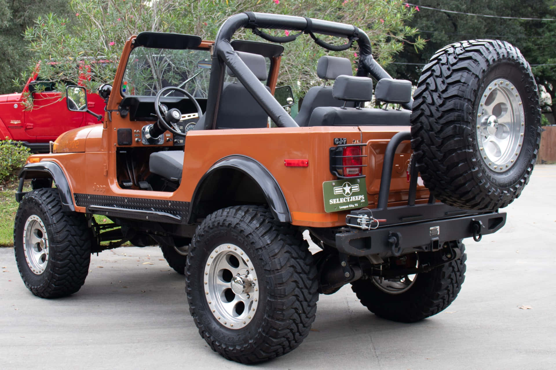 Jeep Cj 桌布 壁紙 — 一輛堅固的橙色Jeep Wrangler，配備超大輪胎和後置備胎，停泊在綠樹環繞的混凝土車道上。