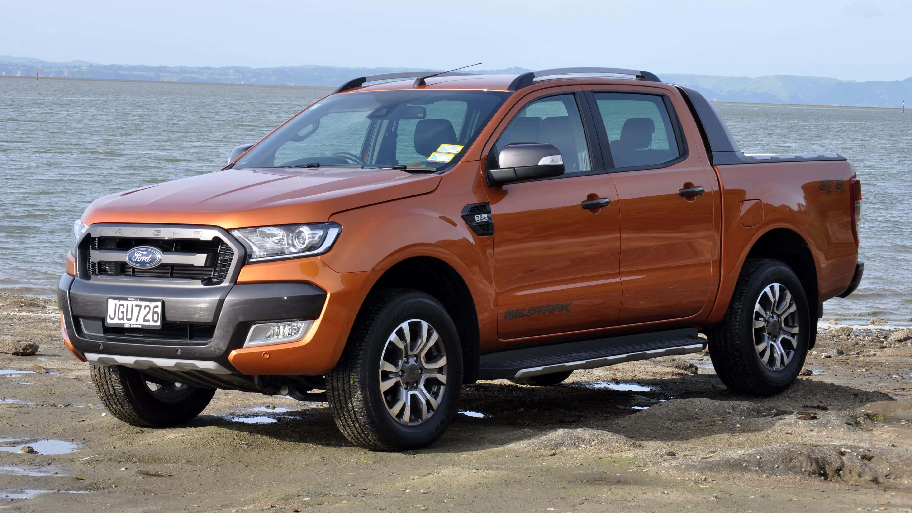 Ford Ranger 桌布 壁紙 — 一輛堅固的橙色福特 Ranger  pickup 貨車停泊在泥濘的岸邊，平靜的水面和遠處的山丘在朦朧的天空下。