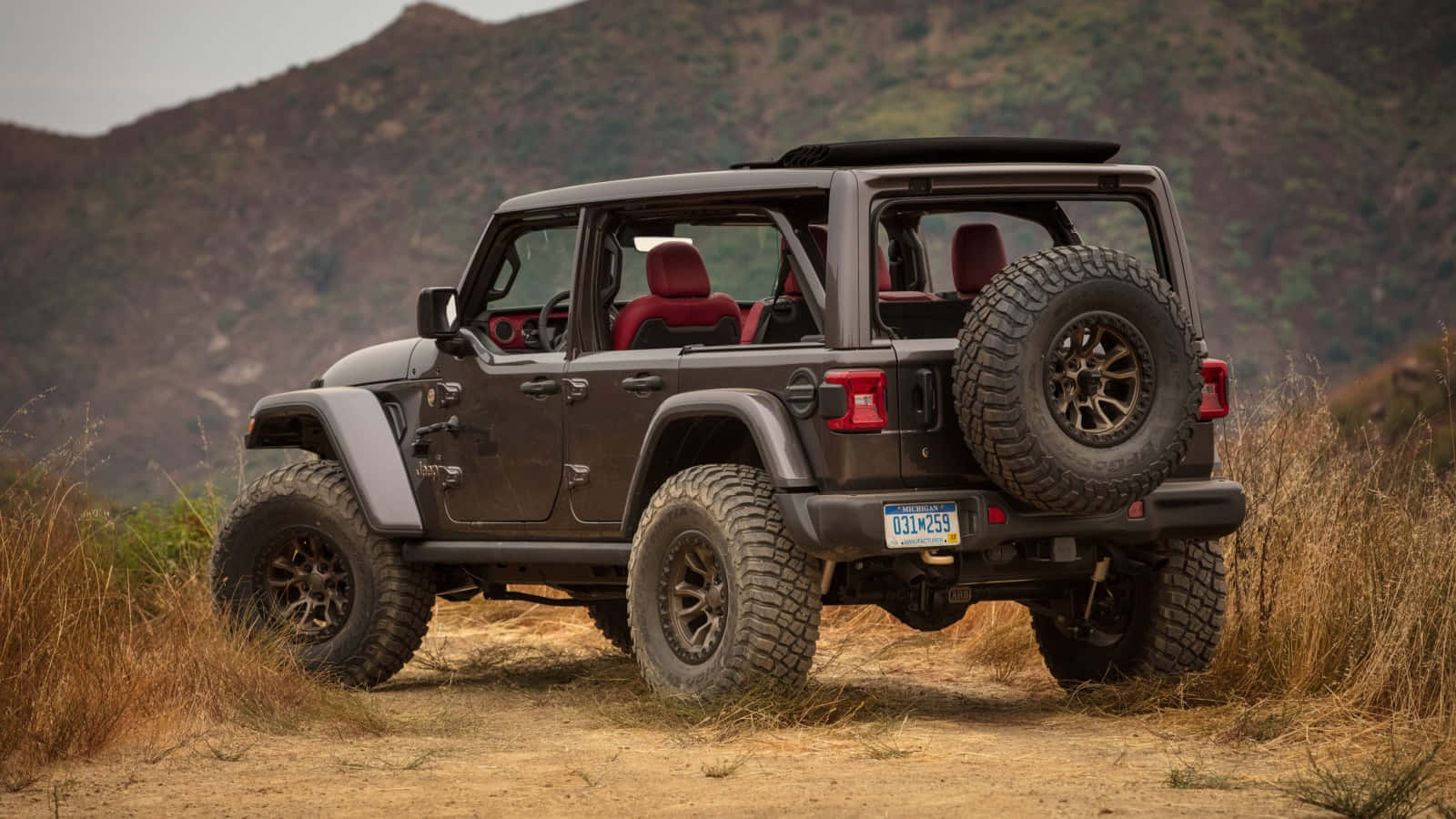 Jeep Wrangler 桌布 壁紙 — 一輛堅固的深棕色Jeep Wrangler，配備紅色內裝和後置備用輪胎，在陰天下行駛於乾燥的草坡。