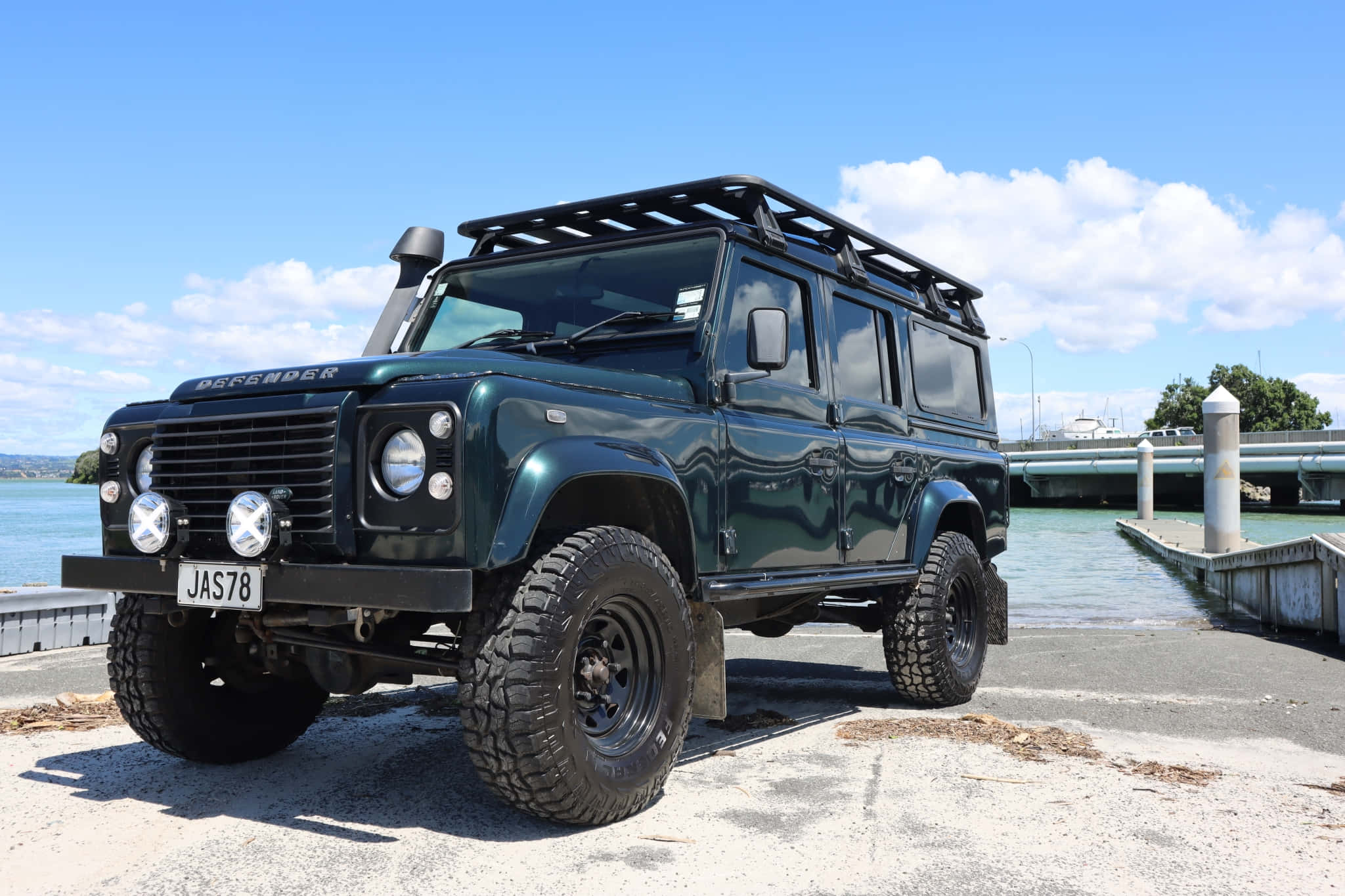 Land Rover Defender 桌布 壁紙 — 一輛堅固的深綠色 Land Rover Defender，配備車頂架和超大輪胎，停泊在混凝土碼頭上，旁邊是平靜的水面，上方是藍天白雲。