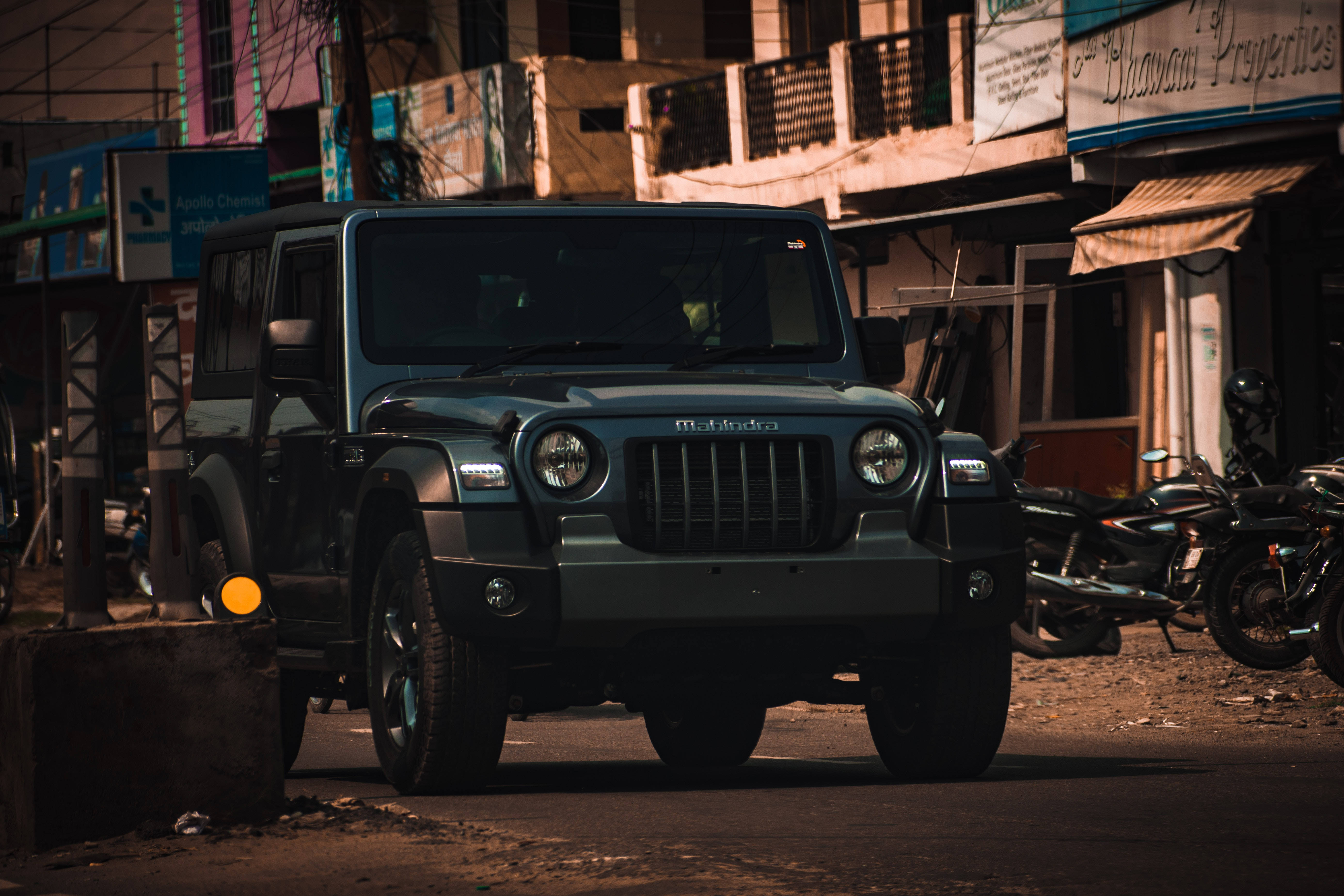 汽車 桌布 壁紙 — 一輛堅固的深綠色Mahindra Thar SUV停泊在塵土飛揚的城市街道上，伴隨著機車和色彩繽紛的建築物，沐浴在溫暖的金色陽光中。