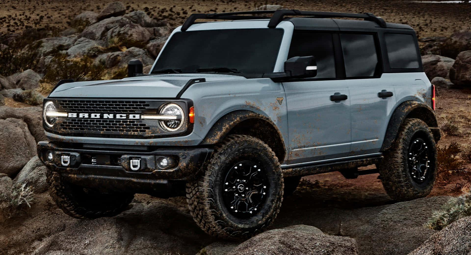 福特野馬越野車 桌布 壁紙 — 一輛堅固的淺藍色福特Bronco SUV，輪胎沾滿泥濘，在明亮的日光下穿越岩石地形。