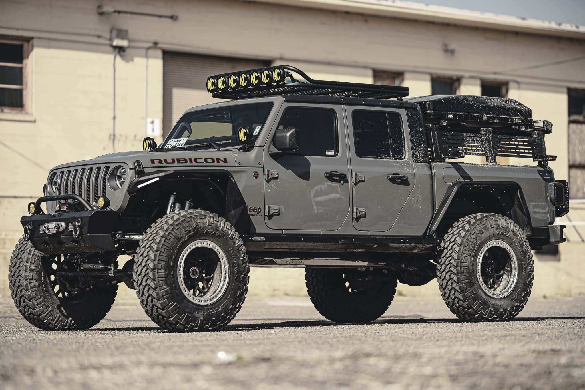 Off-road Car 桌布 壁紙 — 一輛堅固的灰色Jeep Rubicon，配備超大輪胎和車頂燈，停在倉庫前的碎石地上。