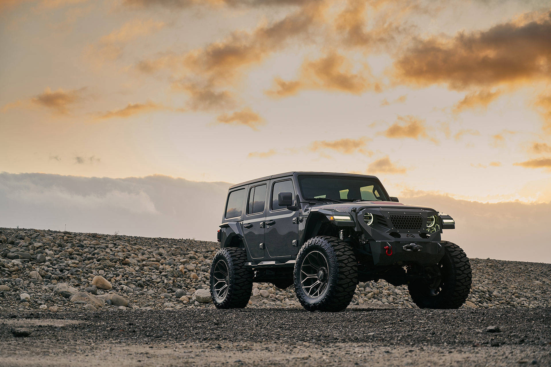 黑色吉普車 桌布 壁紙 — 一輛堅固的灰色Jeep Wrangler配備超大輪胎，在戲劇性的日落天空下矗立在岩石地形上。