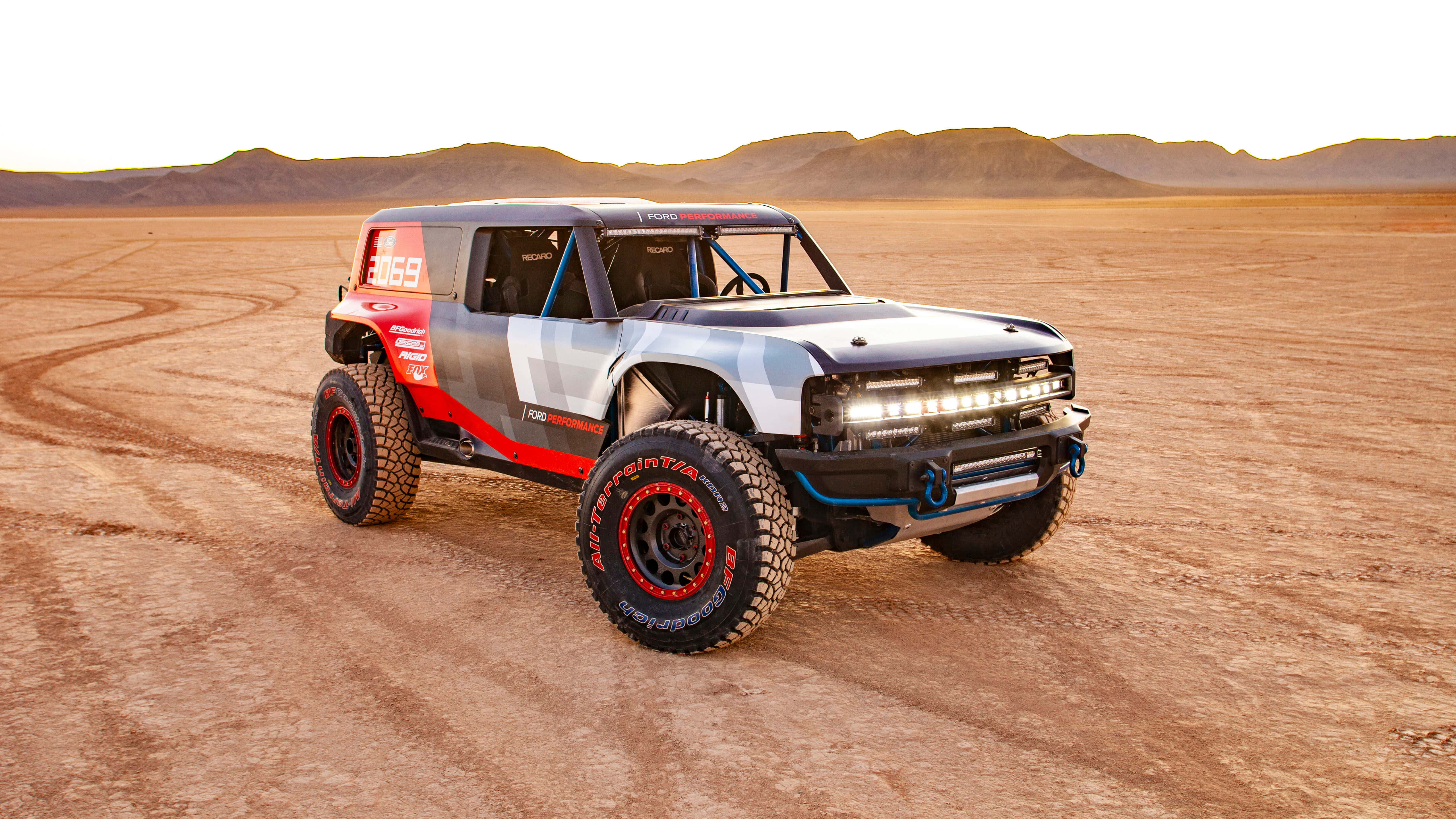 Ford Bronco 桌布 壁紙 — 一輛堅固的福特Bronco賽車卡車，帶有紅色點綴和超大輪胎，在日落時分疾馳穿越廣闊的沙漠景觀。