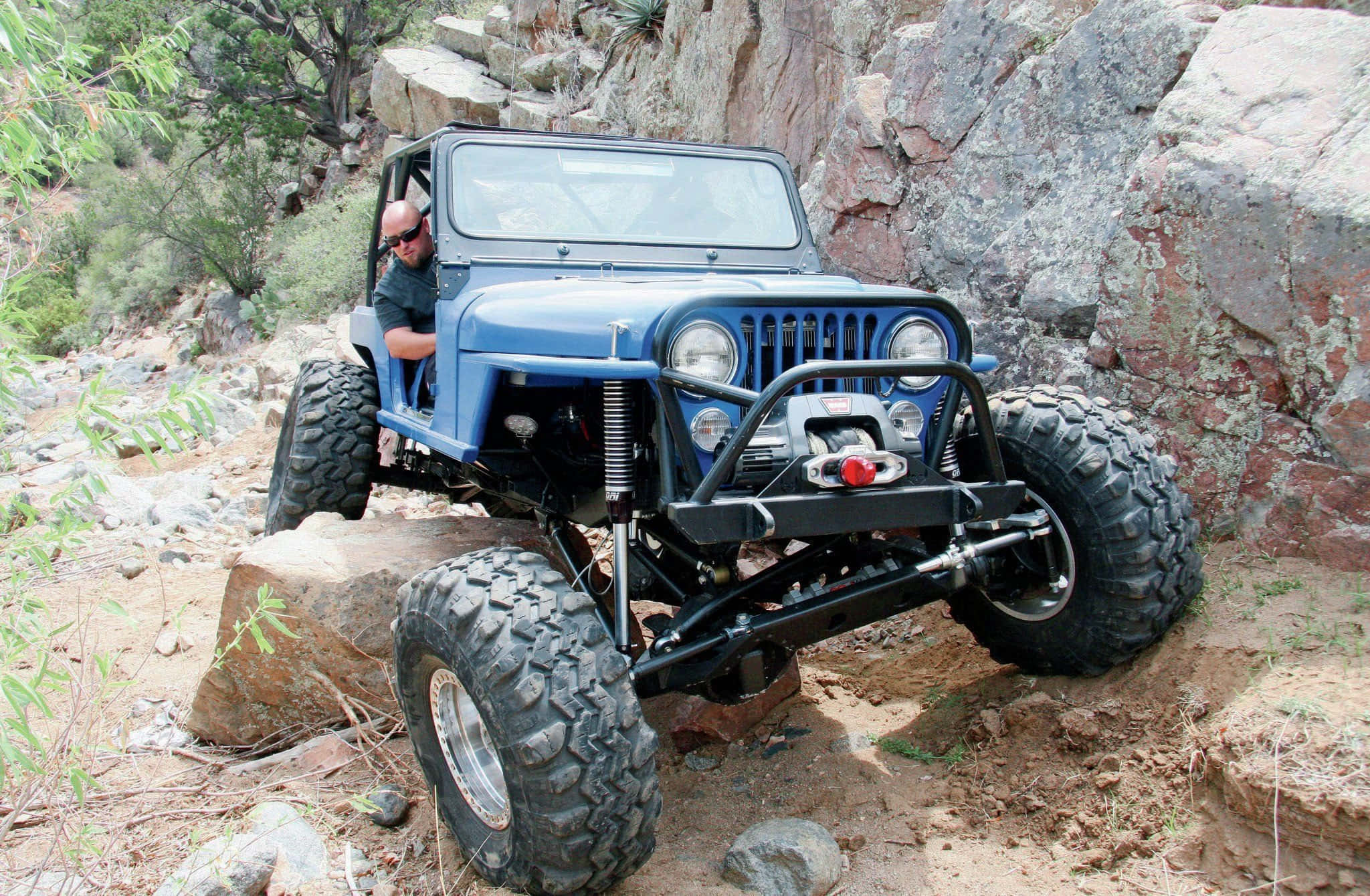 Jeep Cj 桌布 壁紙 — 一輛堅固的藍色Jeep Wrangler配備超大輪胎，攀爬陡峭岩石地形，車內駕駛清晰可見。