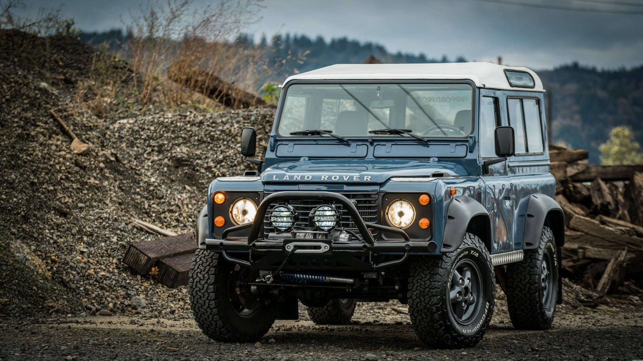 land rover iPhone 桌布 壁紙 — 一輛堅固的藍色Land Rover Defender，白色車頂，停放在碎石堆上，背景是雲天下森林覆蓋的山丘。