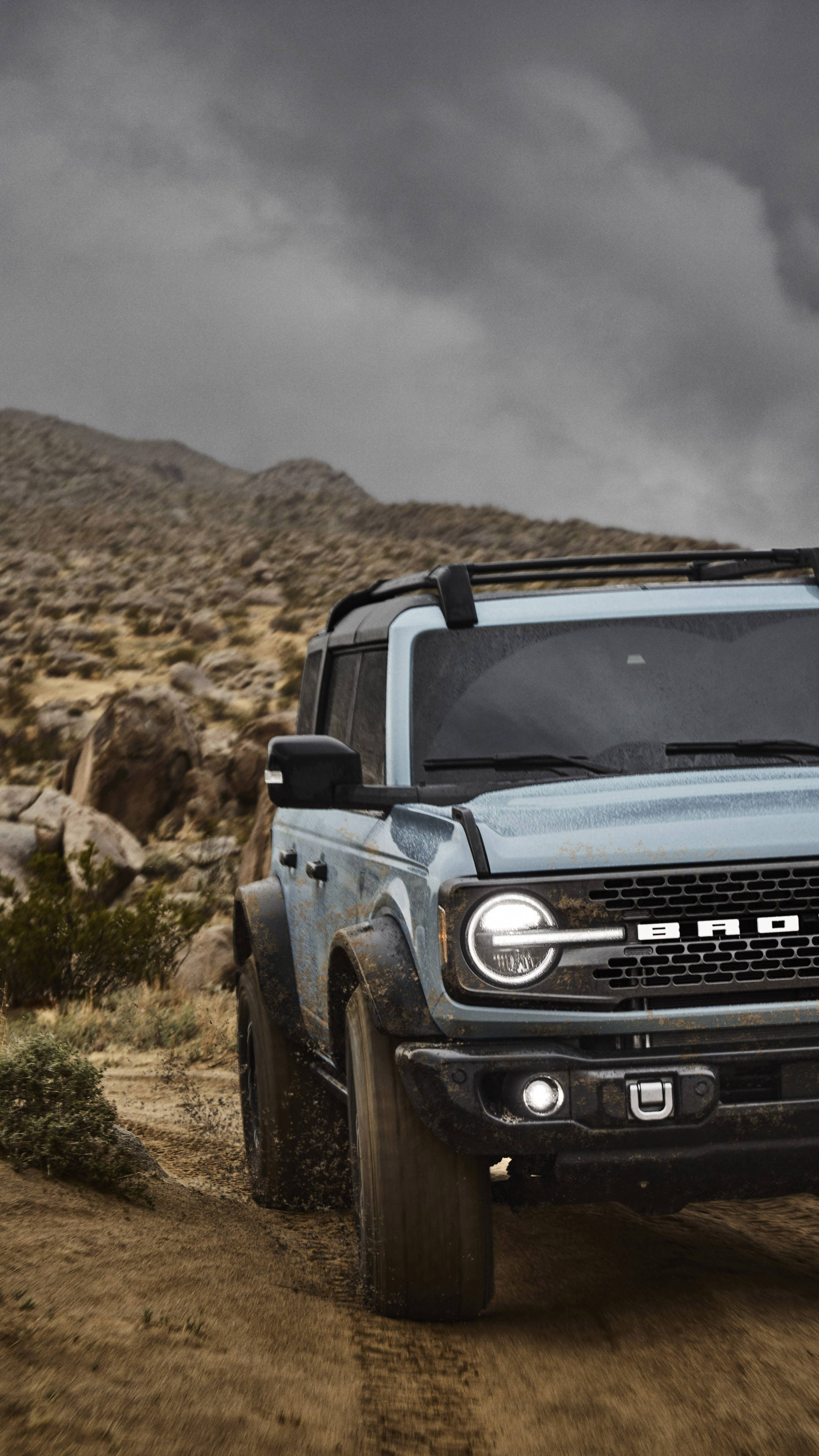 Ford Bronco 桌布 壁紙 — 一輛堅固的藍色福特Bronco在風暴灰天下，背景有岩石丘陵，行駛在塵土飛揚的沙漠小徑上。
