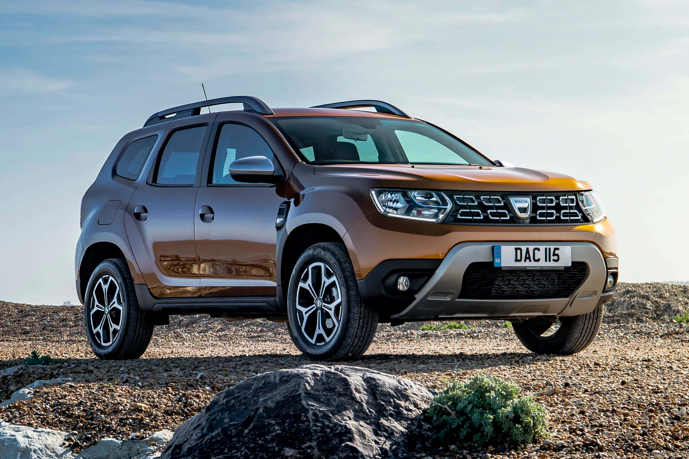Dacia 桌布 壁紙 — 一輛堅固的青銅色Dacia Duster SUV在晴朗的天空下，停放在岩石地形上，附近有稀疏的綠植。