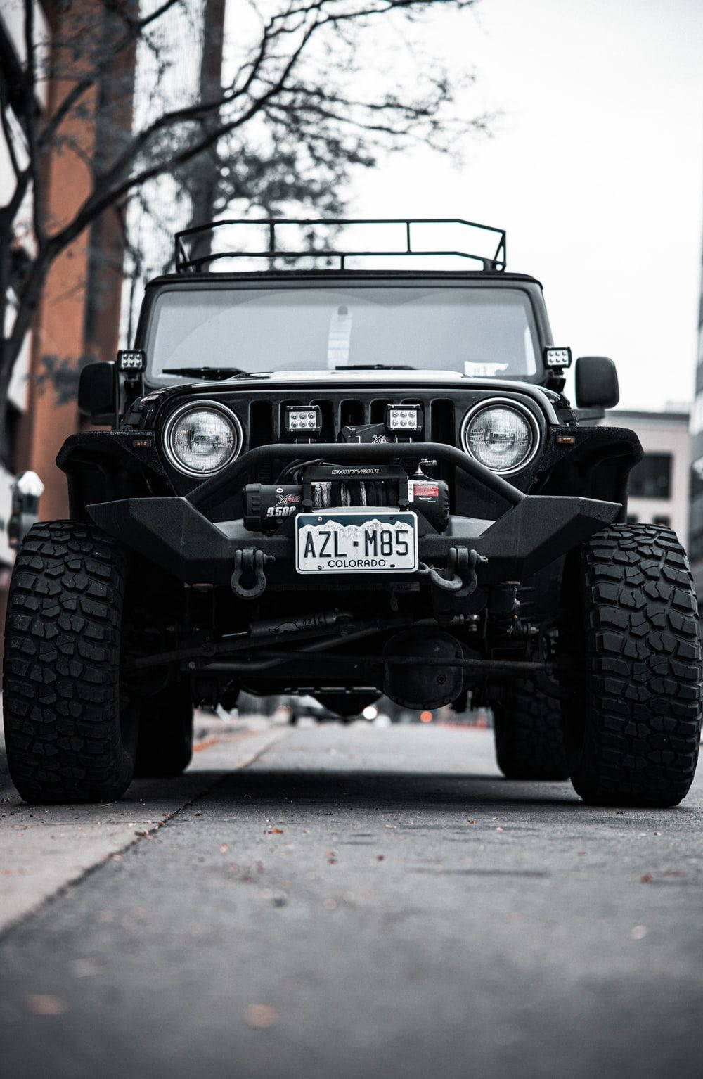 Black Jeep 桌布 壁紙 — 一輛堅固的黑色Jeep Wrangler，配備超大輪胎和車頂行李架，停泊在光禿樹木下的城市街道上。
