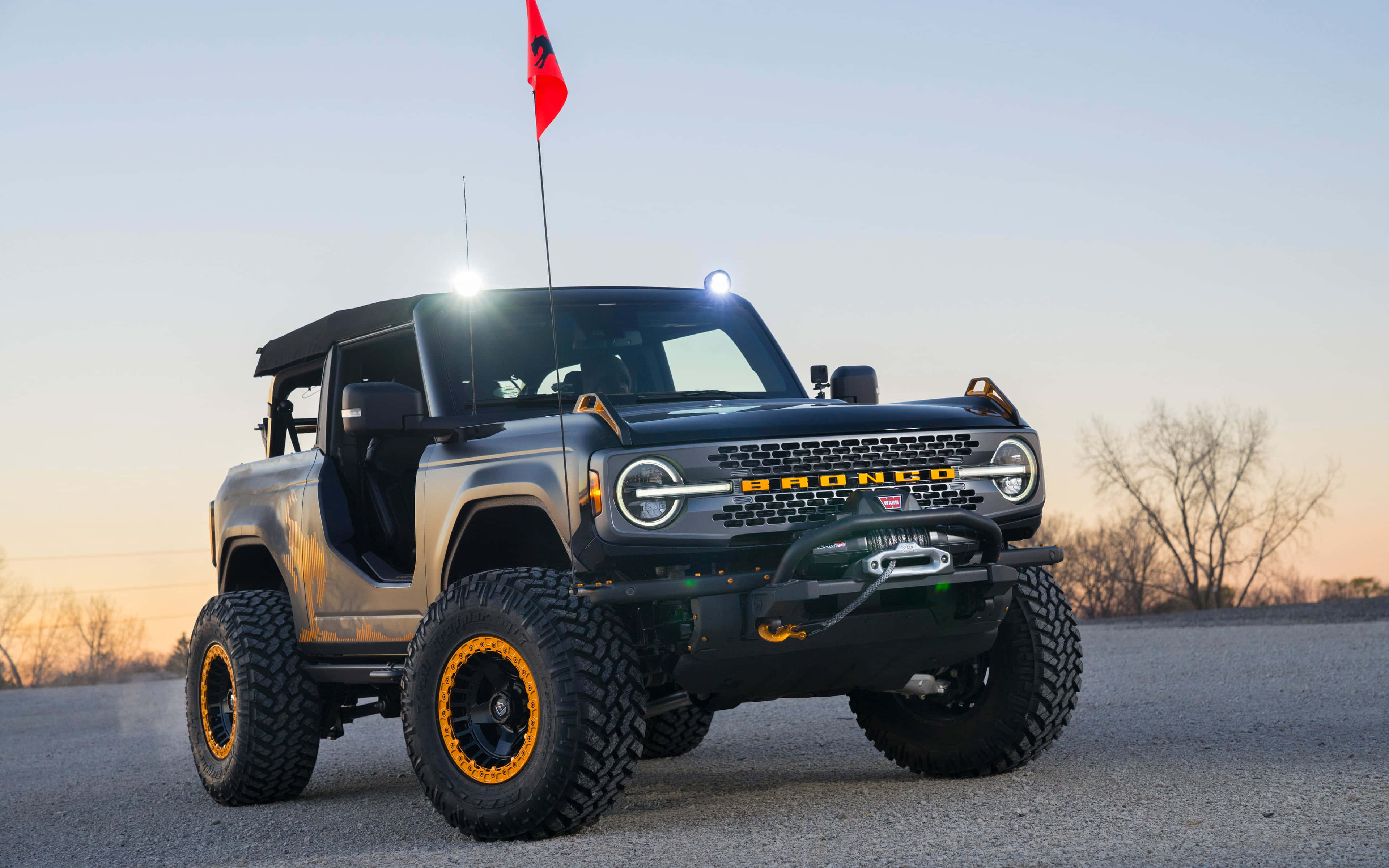 福特野馬越野車 桌布 壁紙 — 一輛堅固的黑色福特Bronco，帶有橙色車輪點綴，在日落時分停在鋪砌的道路上，背景是光禿禿的樹木。
