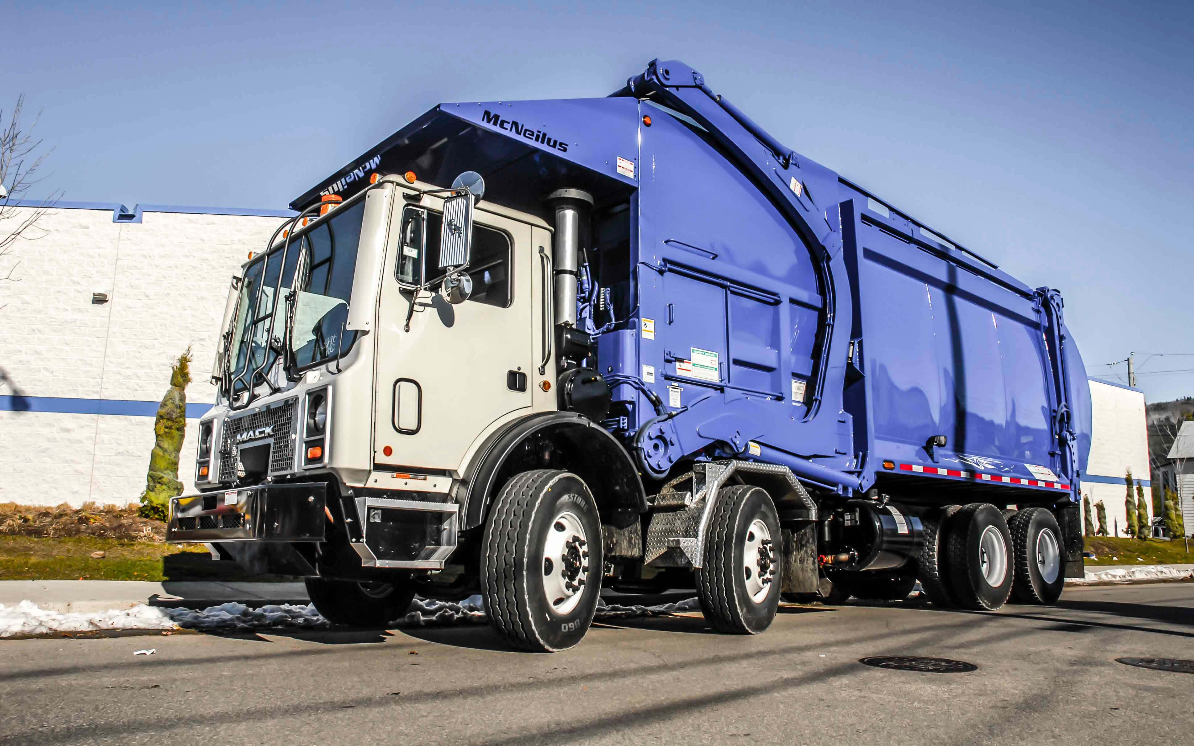 Garbage Truck 桌布 壁紙 — 一輛大型藍白色Mack垃圾車配備McNeilus壓縮機，停在白色建築物附近的鋪砌場地上，天空晴朗湛藍。