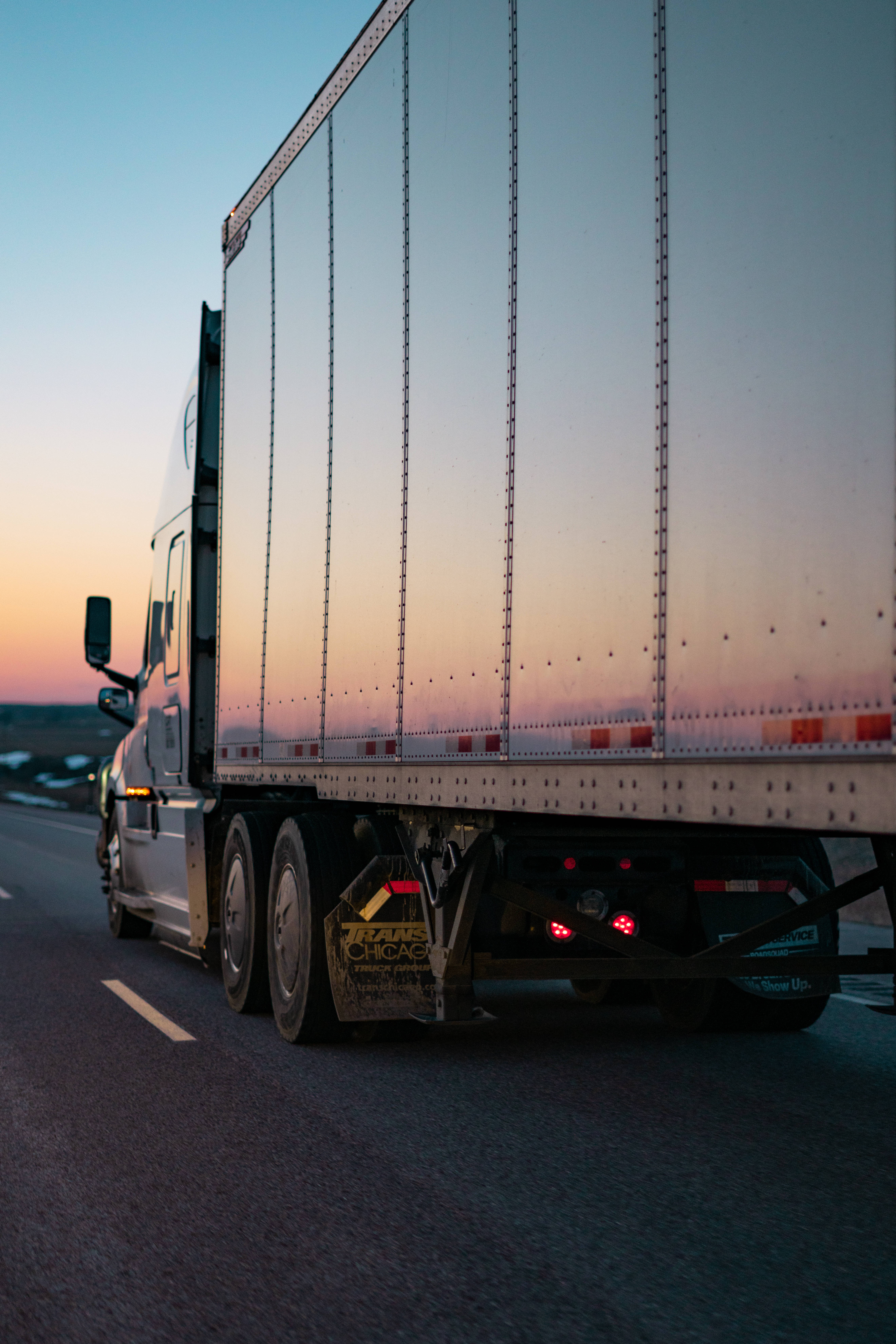 Truck 桌布 壁紙 — 一輛大型銀色半掛卡車在日落時分行駛在高速公路上，溫暖的橙色和粉紅色天空反射在其拖車上。