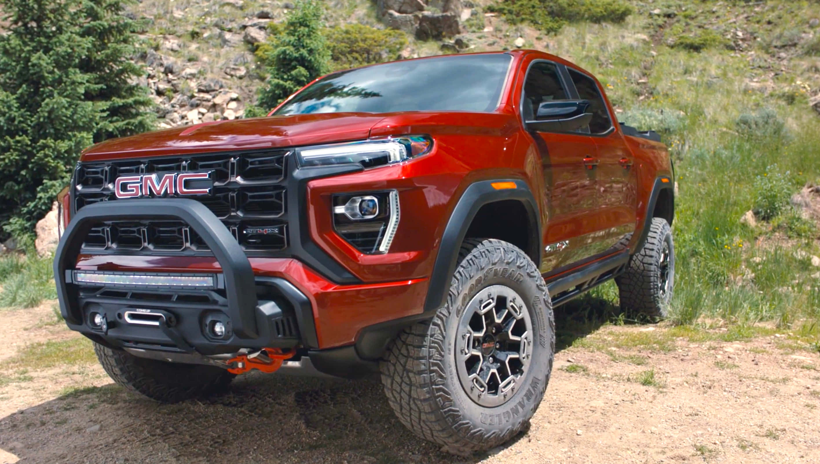 Gmc Canyon 桌布 壁紙 — 一輛大膽的紅色GMC Canyon AT4皮卡車，配備黑色越野保險桿和大輪胎，停放在佈滿綠色灌木的岩石山坡旁邊的泥路上。
