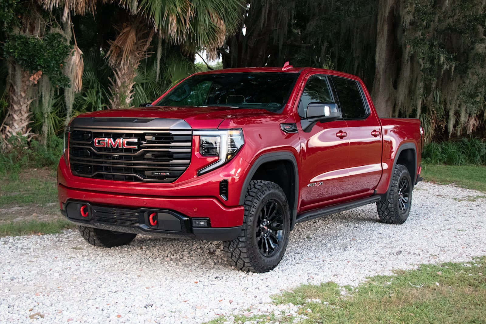 Gmc Sierra 桌布 壁紙 — 一輛大膽的紅色GMC Sierra AT4皮卡車，帶有黑色裝飾和堅固輪胎，停在碎石路上，周圍環繞著茂密的綠植和西班牙苔蘚。