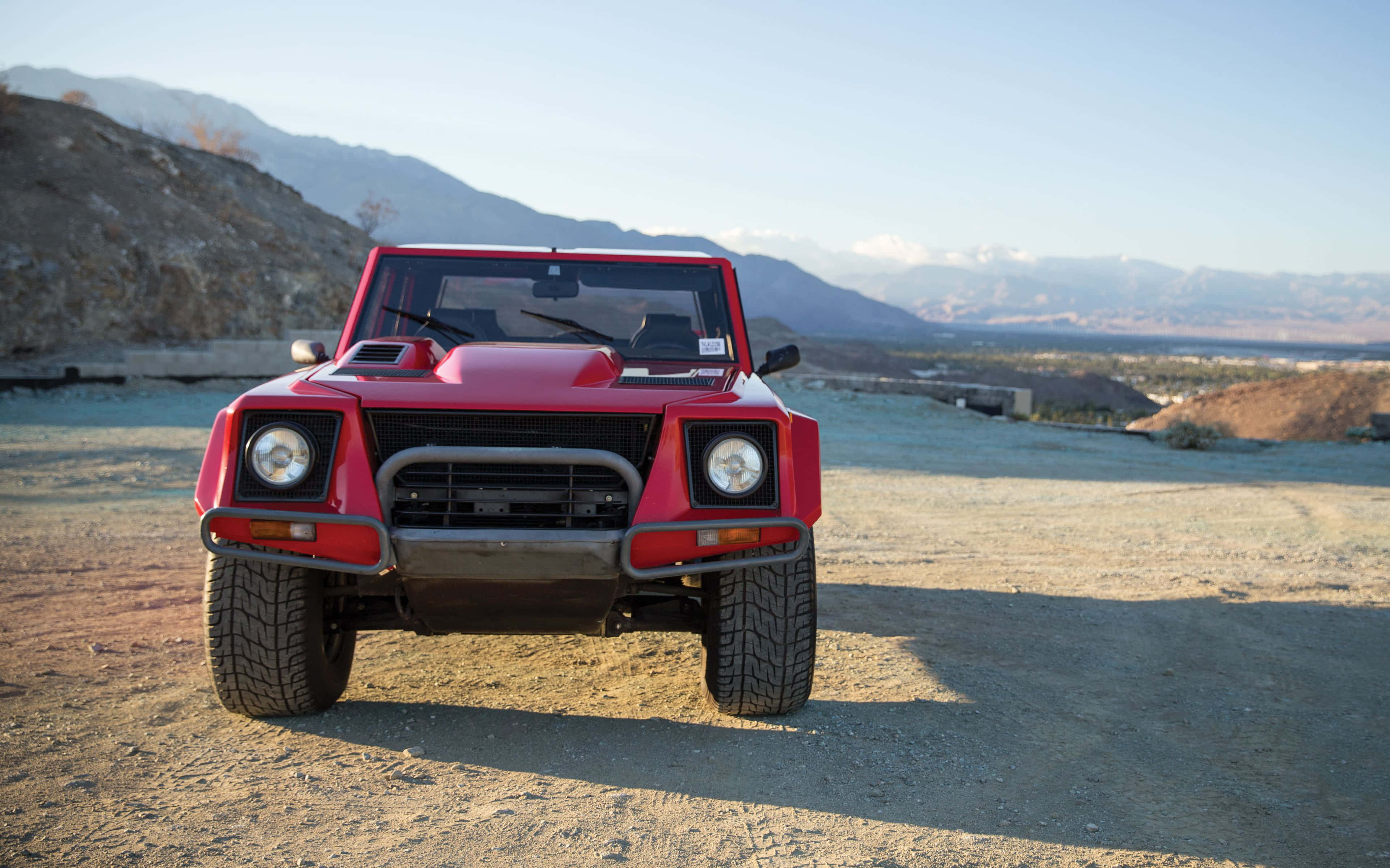 Lamborghini Lm002 桌布 壁紙 — 一輛大膽的紅色藍寶堅尼LM002正面朝向塵土飛揚的地形，背景是崎嶇的山脈和晴朗的天空。