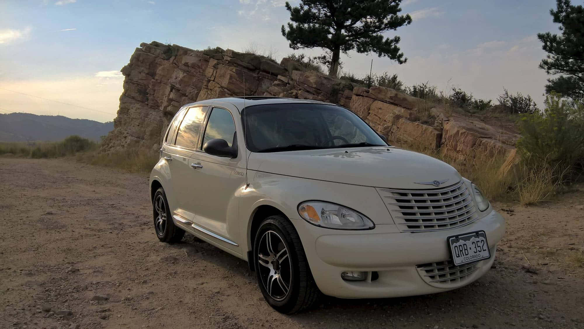 Chrysler Pt Cruiser 桌布 壁紙 — 一輛奶油色的克萊斯勒 PT Cruiser 停在一條塵土飛揚的小徑上，旁邊是岩石峭壁，有一棵孤獨的松樹，背景是柔和的日落天空。
