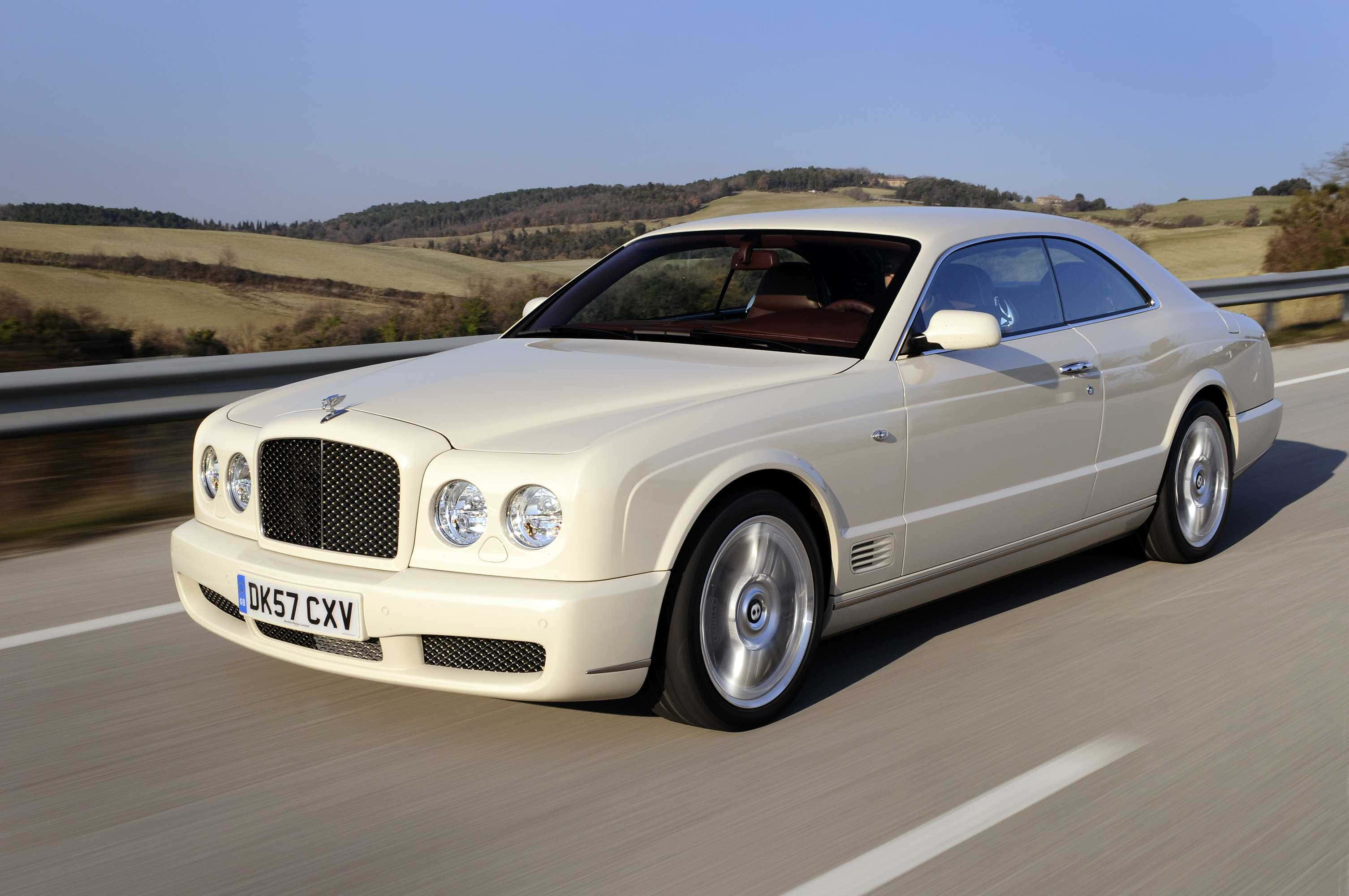 Bentley Brooklands 桌布 壁紙 — 一輛奶油色的賓利 Continental GT 在陽光明媚的高速公路上行駛，背景是連綿起伏的綠色山丘。