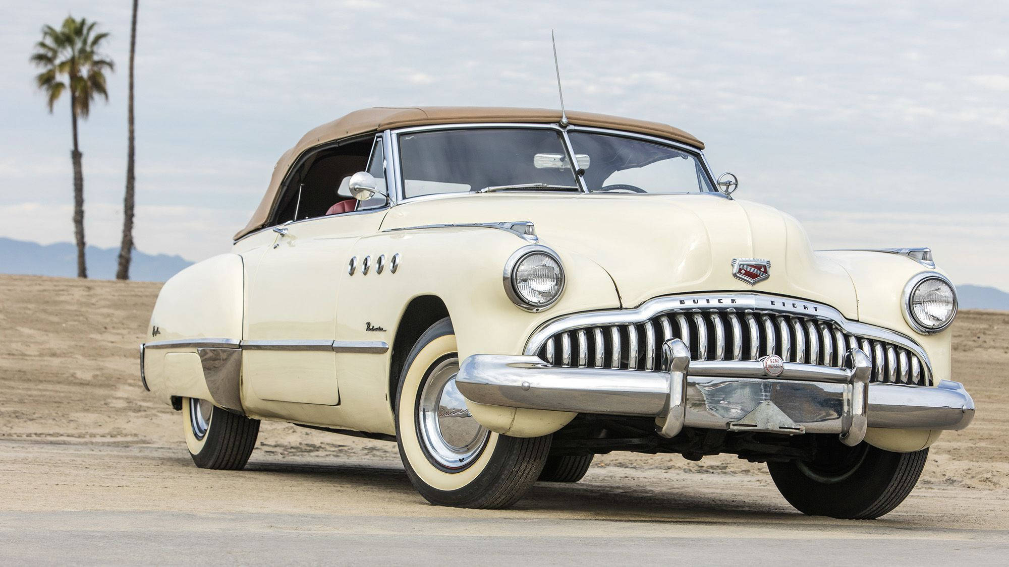 Buick 桌布 壁紙 — 一輛奶油色經典敞篷車配米色軟頂，停泊在沙漠公路上，天空多雲，有棕櫚樹和遠山。