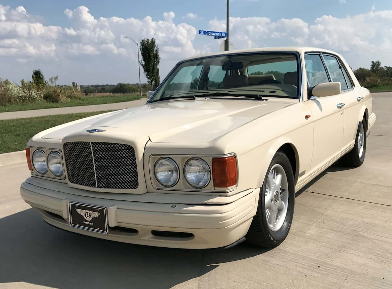 Bentley Eight 桌布 壁紙 — 一輛奶油色賓利轎車在明亮的日光下閃閃發光，停在混凝土停車場上，背景有藍色路牌和綠化植物。