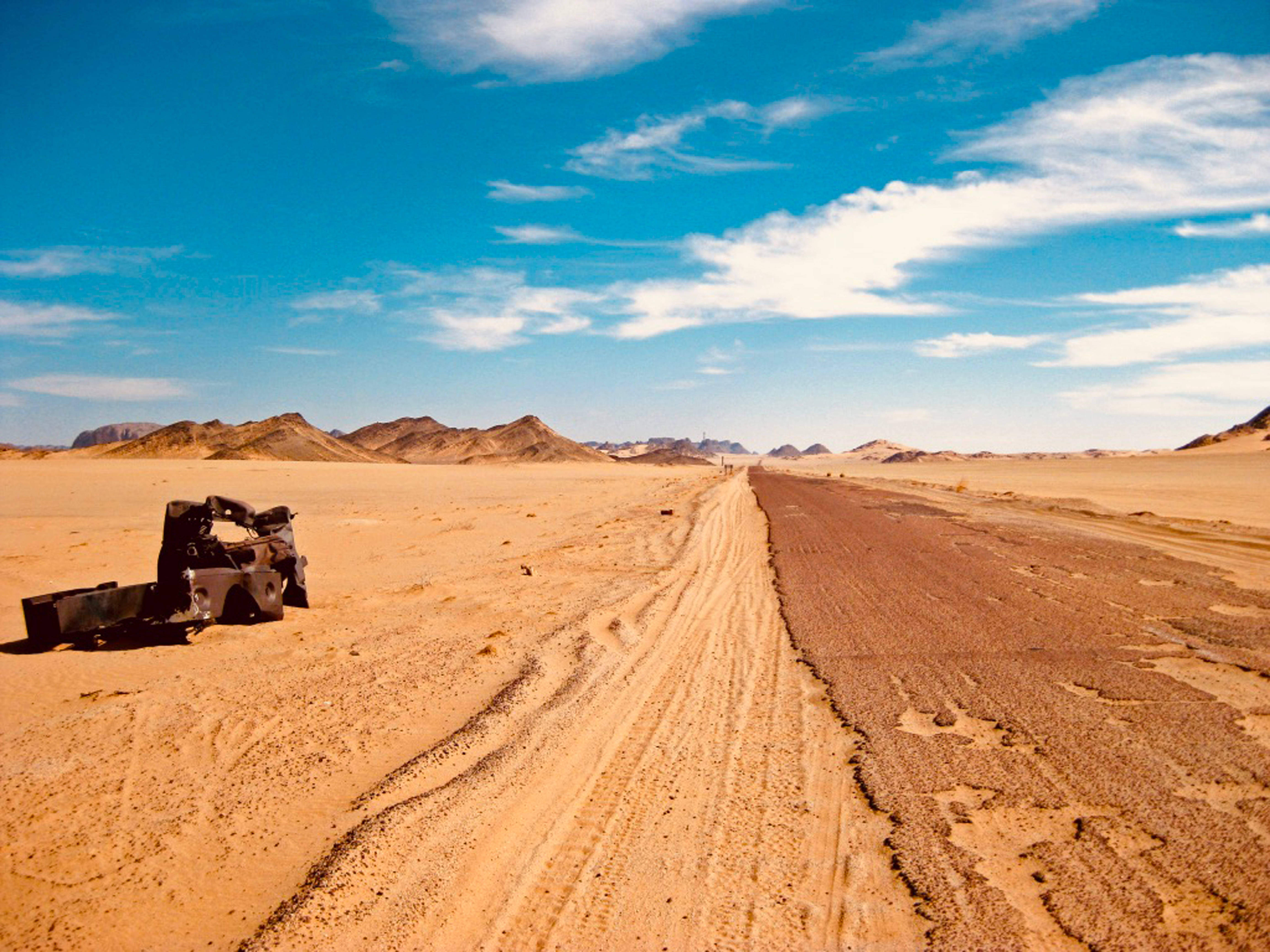 Algeria 桌布 壁紙 — 一輛孤獨的廢棄車輛停泊在蜿蜒的沙漠公路旁，上方是廣闊的藍天，點綴著幾朵白雲。