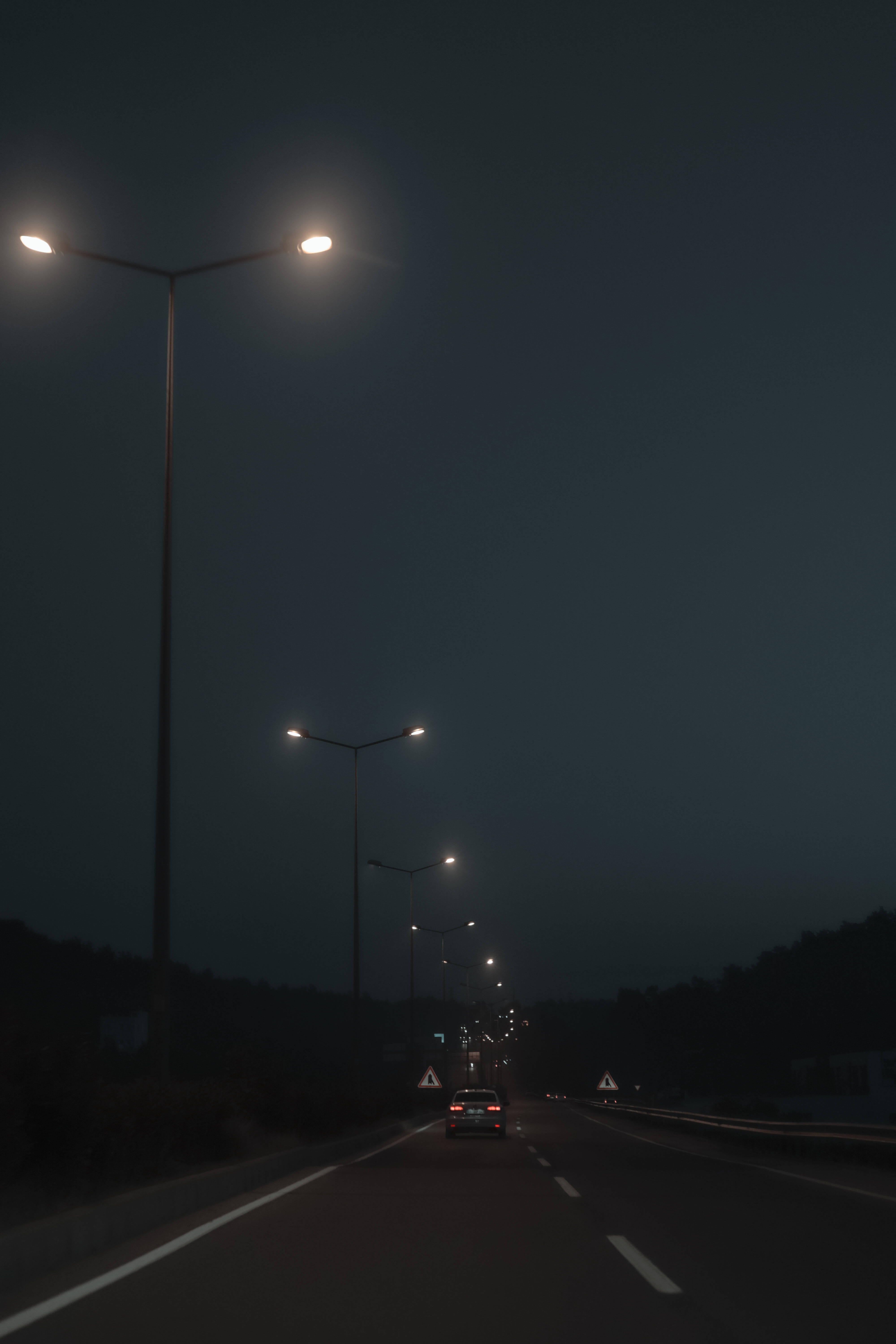 Dim 桌布 壁紙 — 一輛孤獨的汽車在夜間行駛在一條昏暗的高速公路上，下方是發光的街燈，路邊排列著黑暗的樹木。