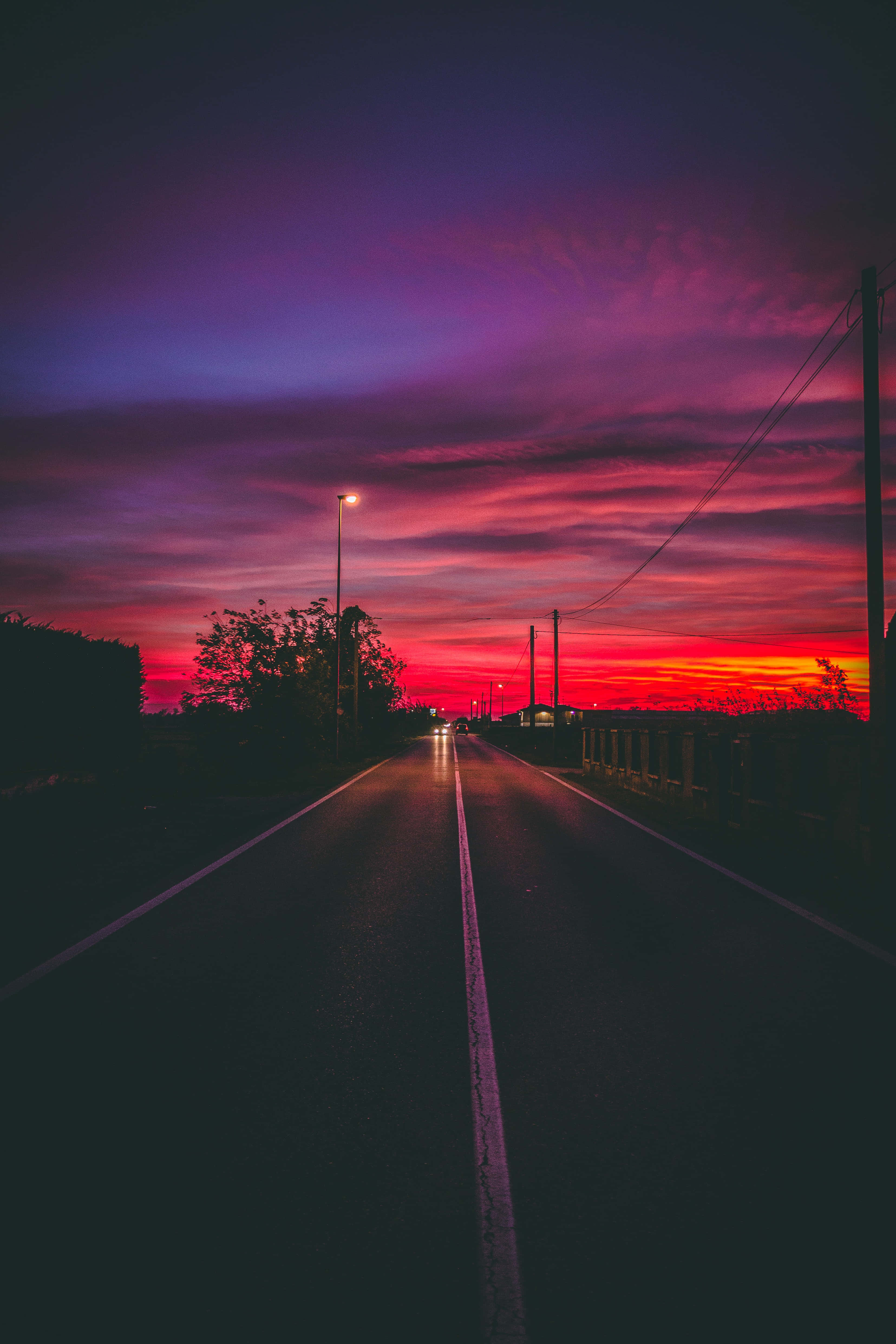 Road 桌布 壁紙 — 一輛孤獨的汽車在戲劇性的紫色和紅色日落天空下行駛在一條黑暗的道路上。
