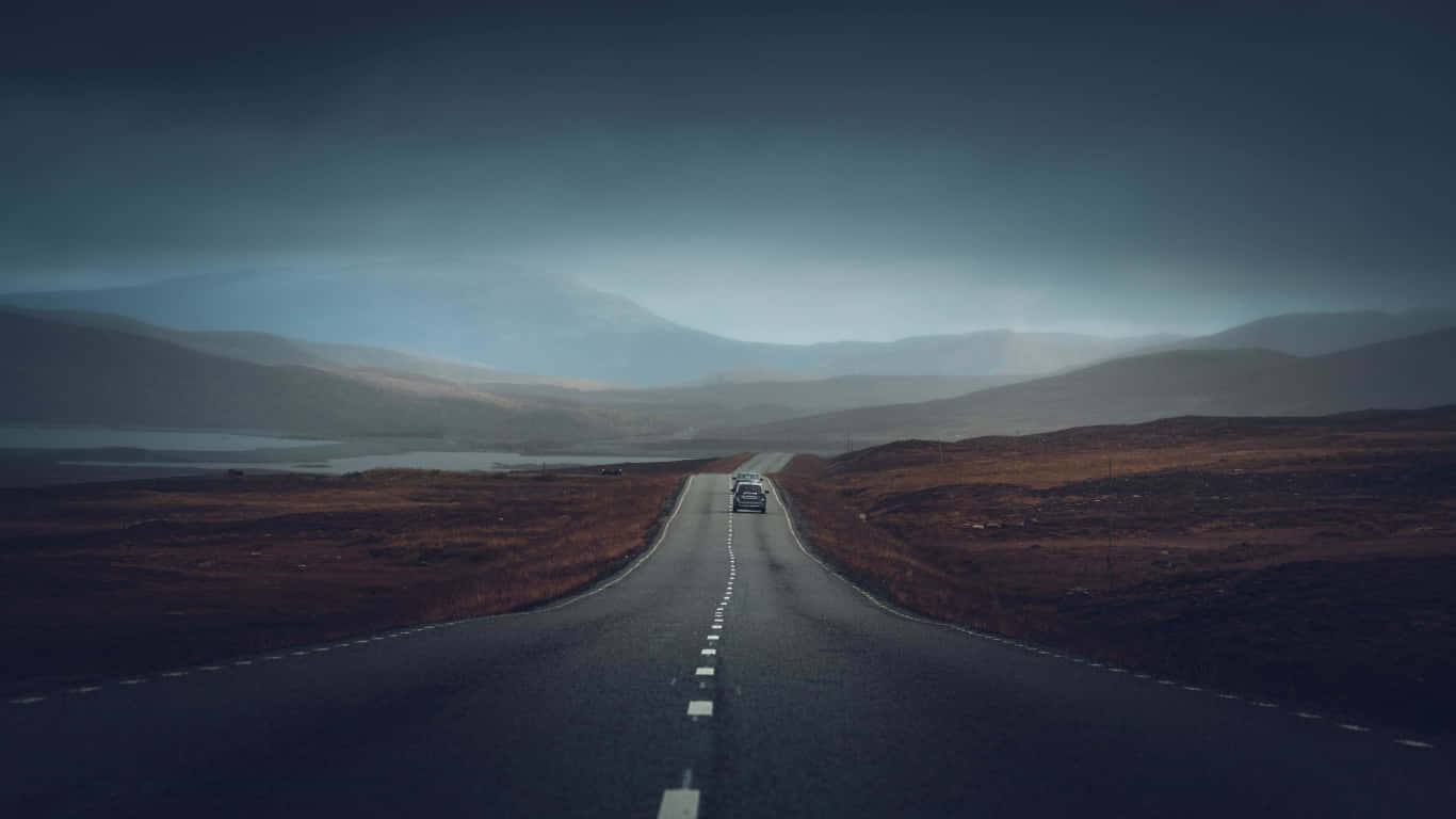 Road 桌布 壁紙 — 一輛孤獨的汽車在陰暗多雲的天空下，沿著蜿蜒的道路穿過霧氣瀰漫的山脈行駛。