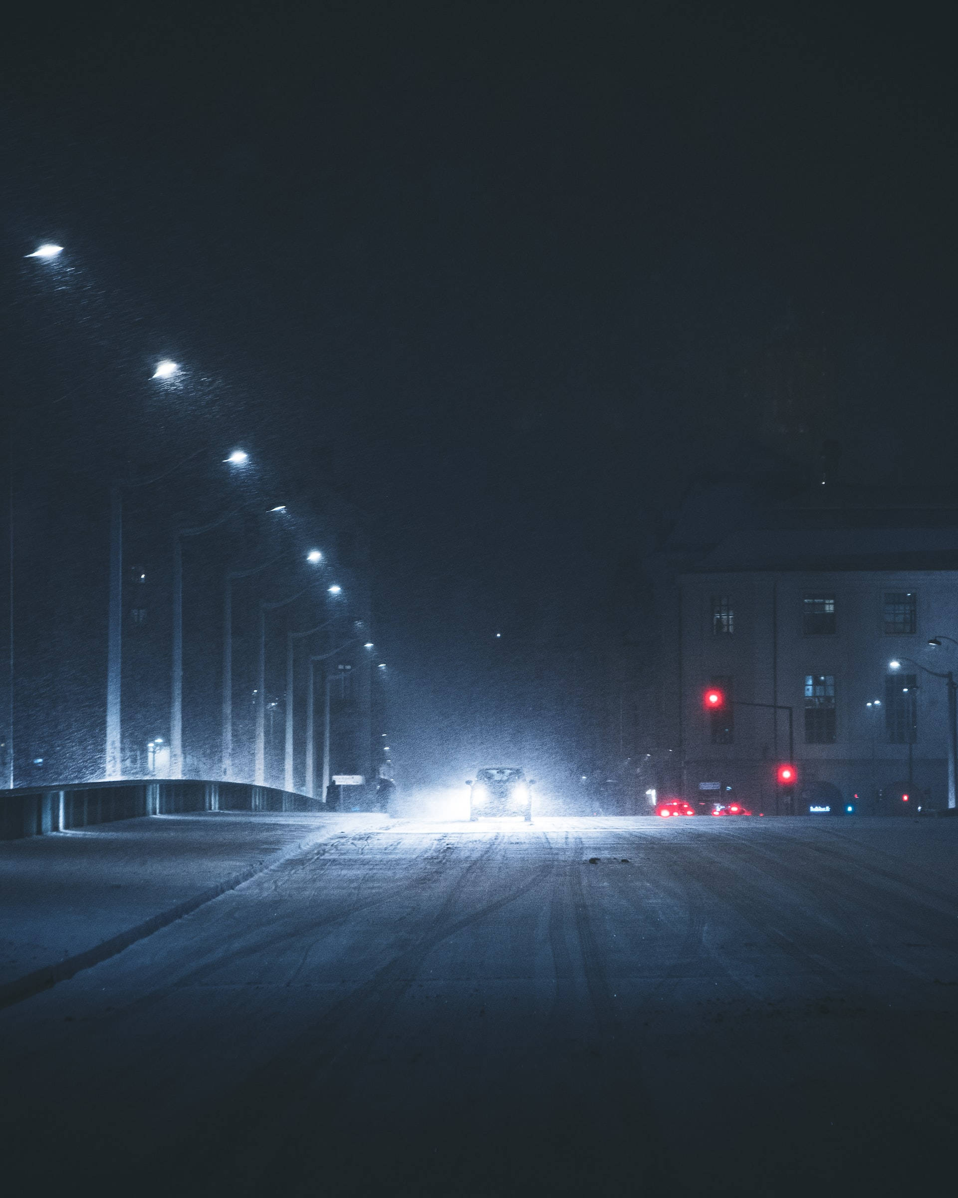 雪 桌布 壁紙 — 一輛孤獨的汽車車頭燈穿透積雪的黑暗，行駛在一條空蕩蕩的市區街道上，兩旁是路燈和紅色交通號誌。