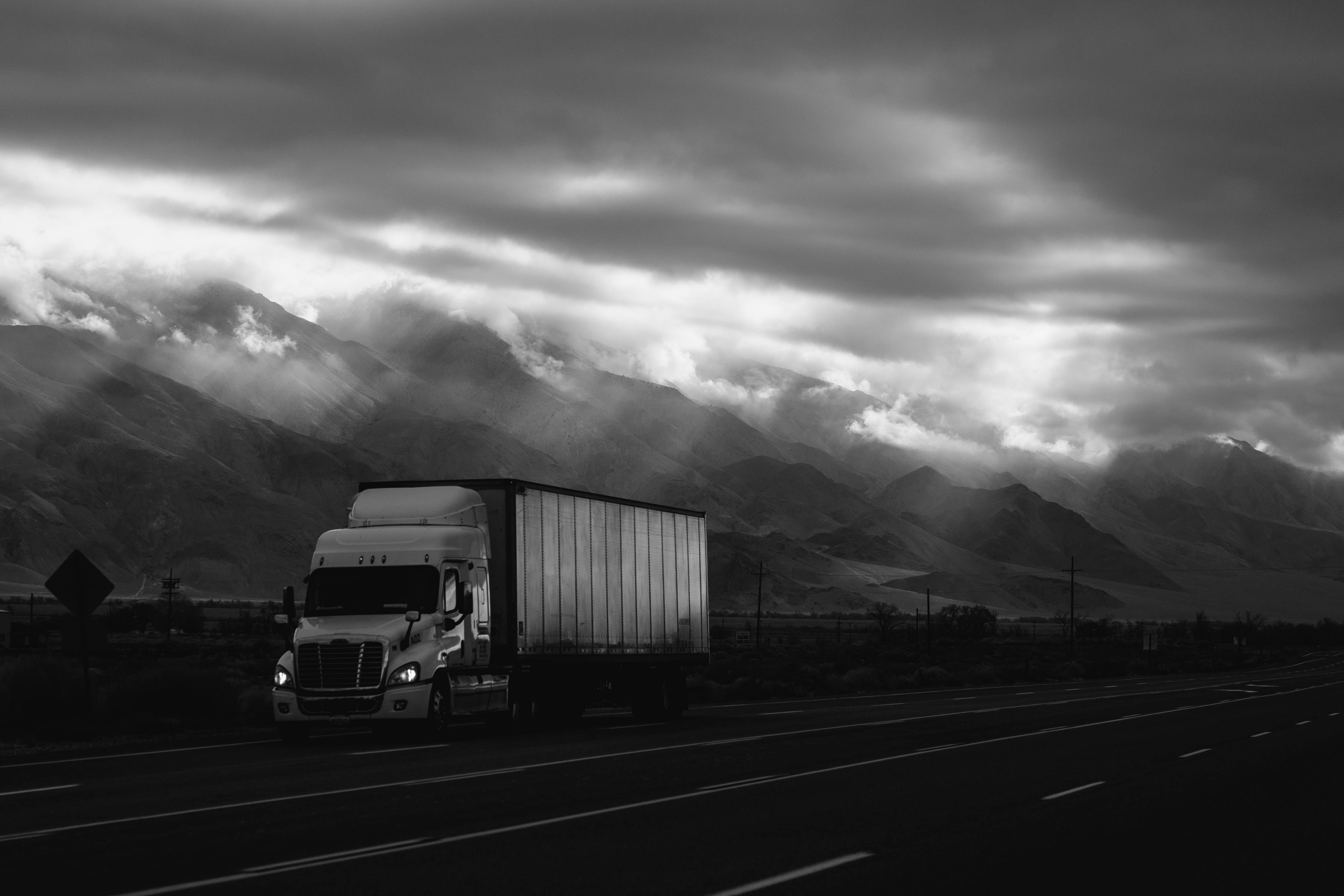 Truck 桌布 壁紙 — 一輛孤獨的貨櫃車在單色調的戲劇性暴風雲和 towering 山脈下行駛在沙漠公路上。