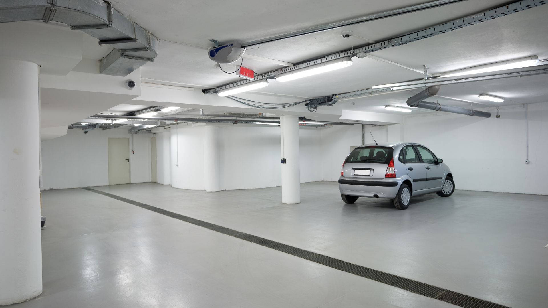 Garage 桌布 壁紙 — 一輛孤獨的銀色汽車停放在一個明亮空曠的地下停車場，有白色柱子和暴露的天花板管道。