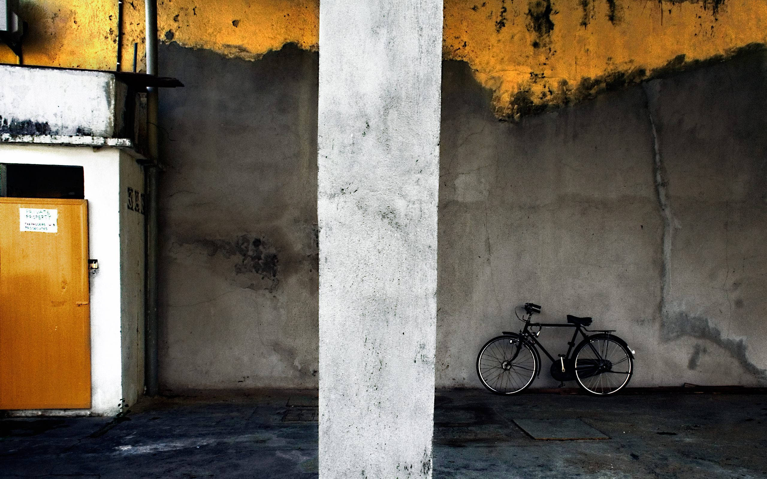 Bicycle 桌布 壁紙 — 一輛孤獨的黑色自行車靠在一面風化的混凝土牆上，上方是城市小巷中大膽的黃色天花板。