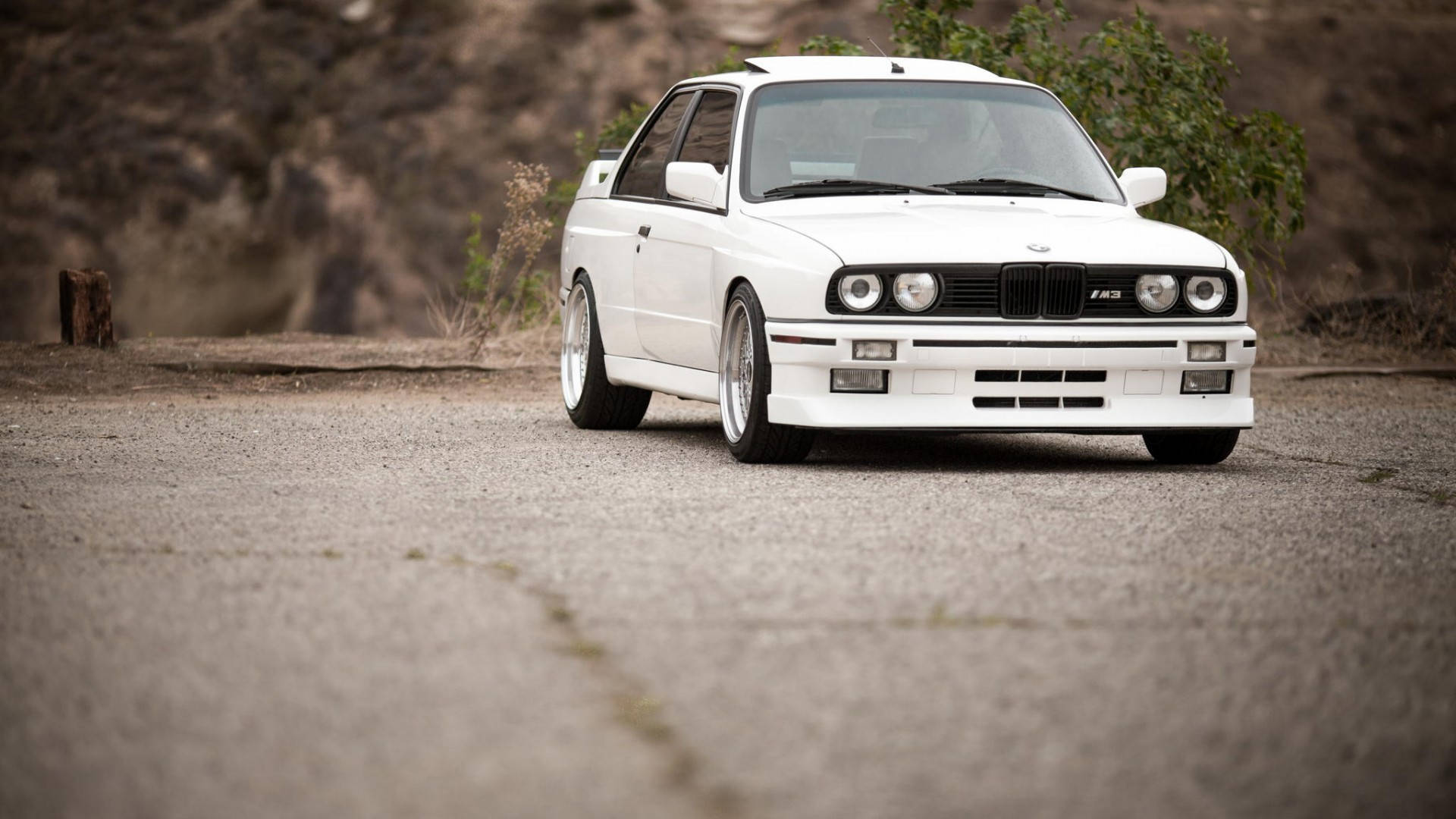 m 桌布 壁紙 — 一輛完美無瑕的白色BMW E30 M3跑車停泊在碎石路上，旁邊是岩石山坡，植被稀疏。