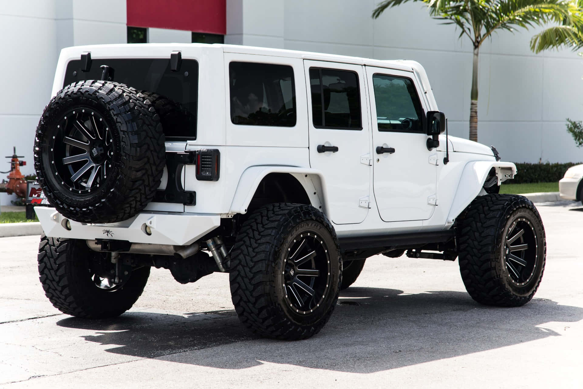 Jeep Wrangler 桌布 壁紙 — 一輛完美無瑕的白色升高Jeep Wrangler，配備超大黑色輪胎和安裝的備用輪胎，停放在陽光明媚的混凝土場地上。