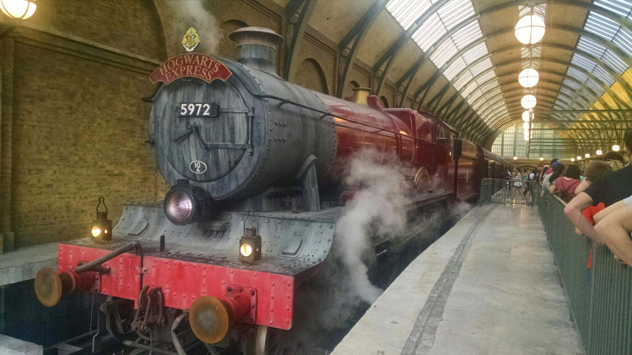The Hogwarts Express Train 桌布 壁紙 — 一輛宏偉的紅色霍格華茲特快蒸汽火車在一個宏偉的拱形車站月台上噴出煙霧，遊客們在觀看。
