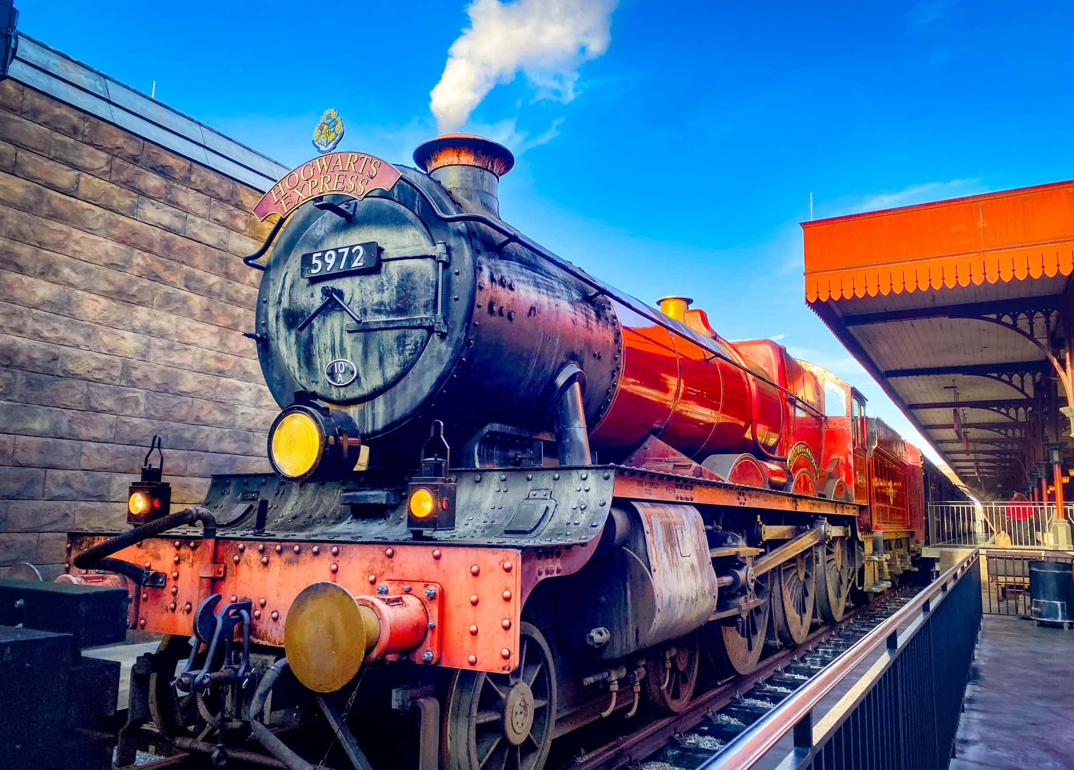 The Hogwarts Express Train 桌布 壁紙 — 一輛宏偉的紅色霍格華茲特快蒸汽火車在晴朗藍天下的車站噴出煙霧。