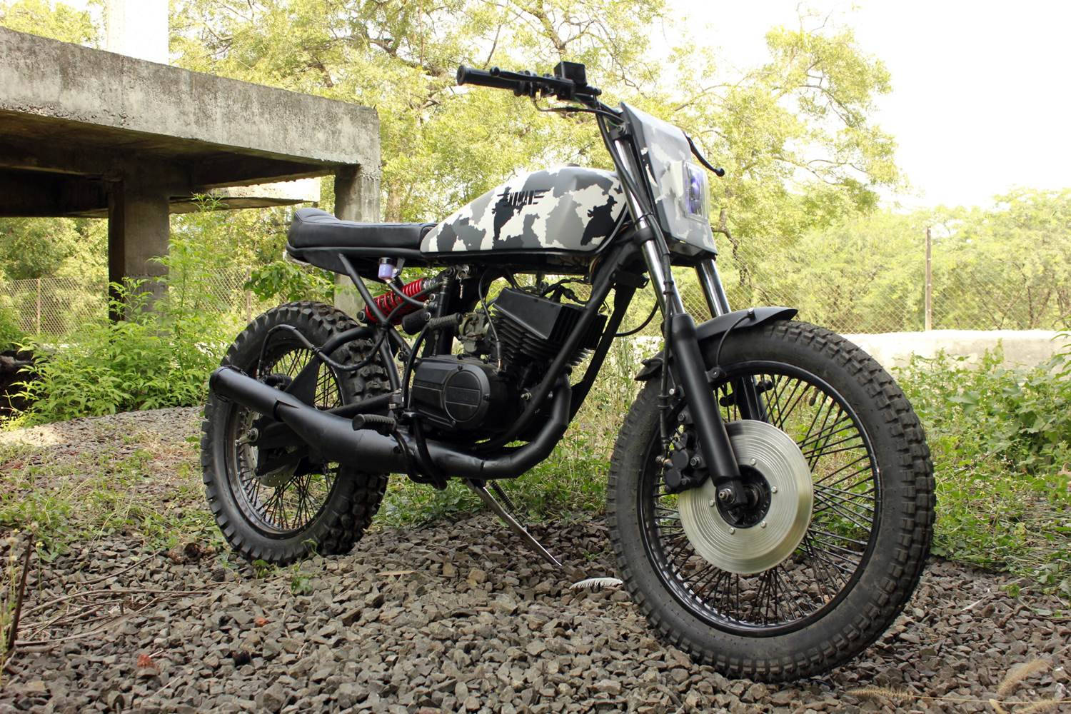 Rx100 桌布 壁紙 — 一輛定制迷彩圖案的摩托車，配備越野輪胎，停在混凝土高架橋下的礫石上，周圍環繞著綠色植物。