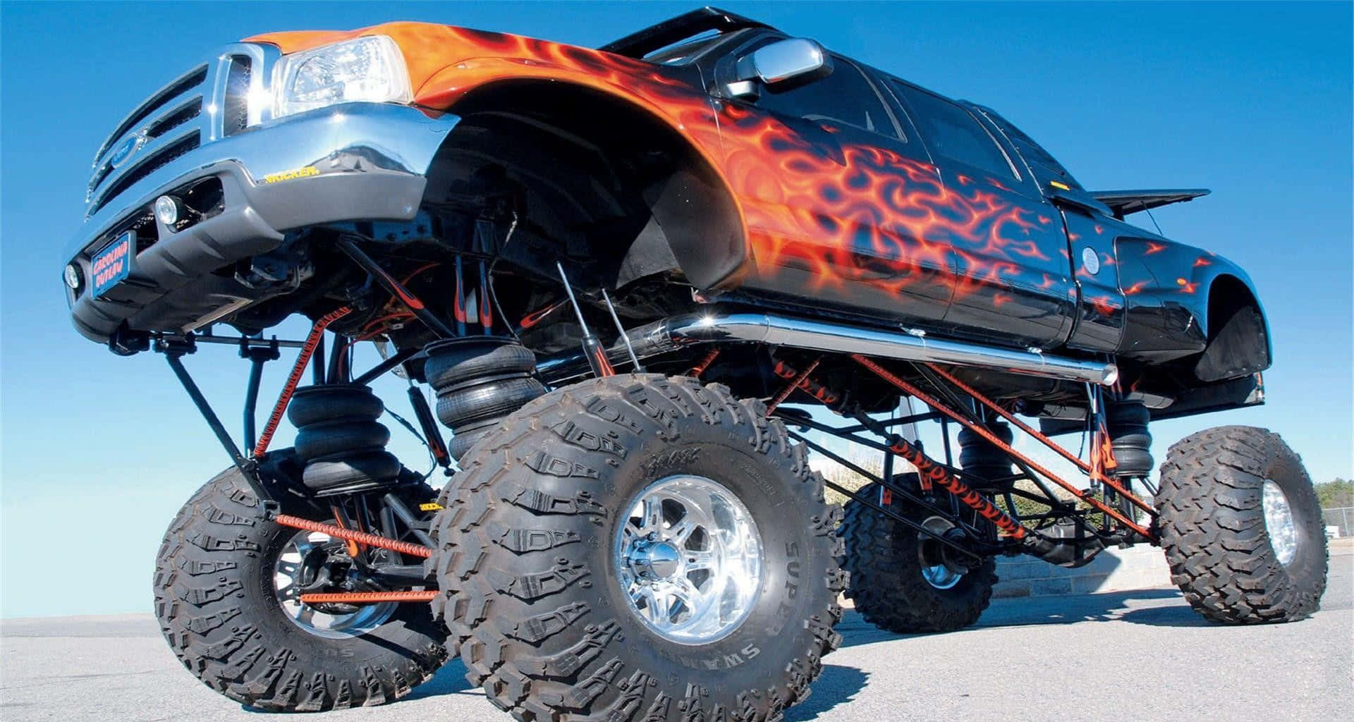 Cool Car 桌布 壁紙 — 一輛定製升高怪獸卡車，帶有火熱橙色火焰貼花和巨大輪胎，在晴朗的藍天下傲然挺立。