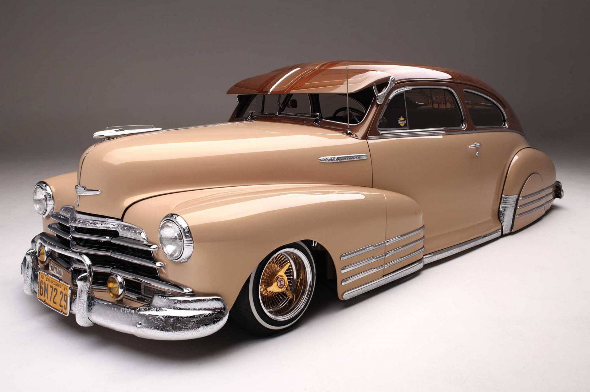 Lowrider 桌布 壁紙 — 一輛定製的低底盤經典車，米色車身和棕色車頂在攝影棚燈光下閃耀，展示鍍鉻裝飾和金色輪圈。