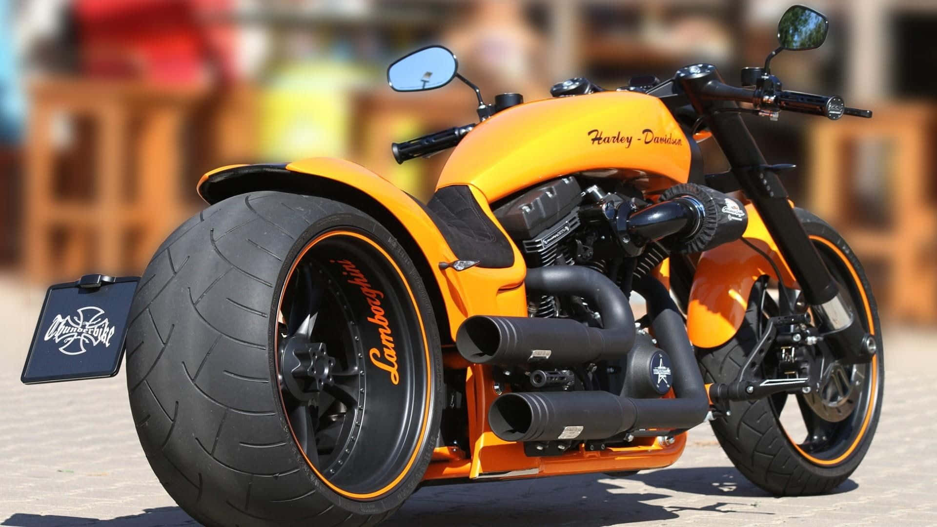 Harley Davidson HD 桌布 壁紙 — 一輛定製的橙色哈雷戴維森摩托車，輪胎上帶有藍寶堅尼品牌標誌，停泊在陽光普照的鋪砌地面上。