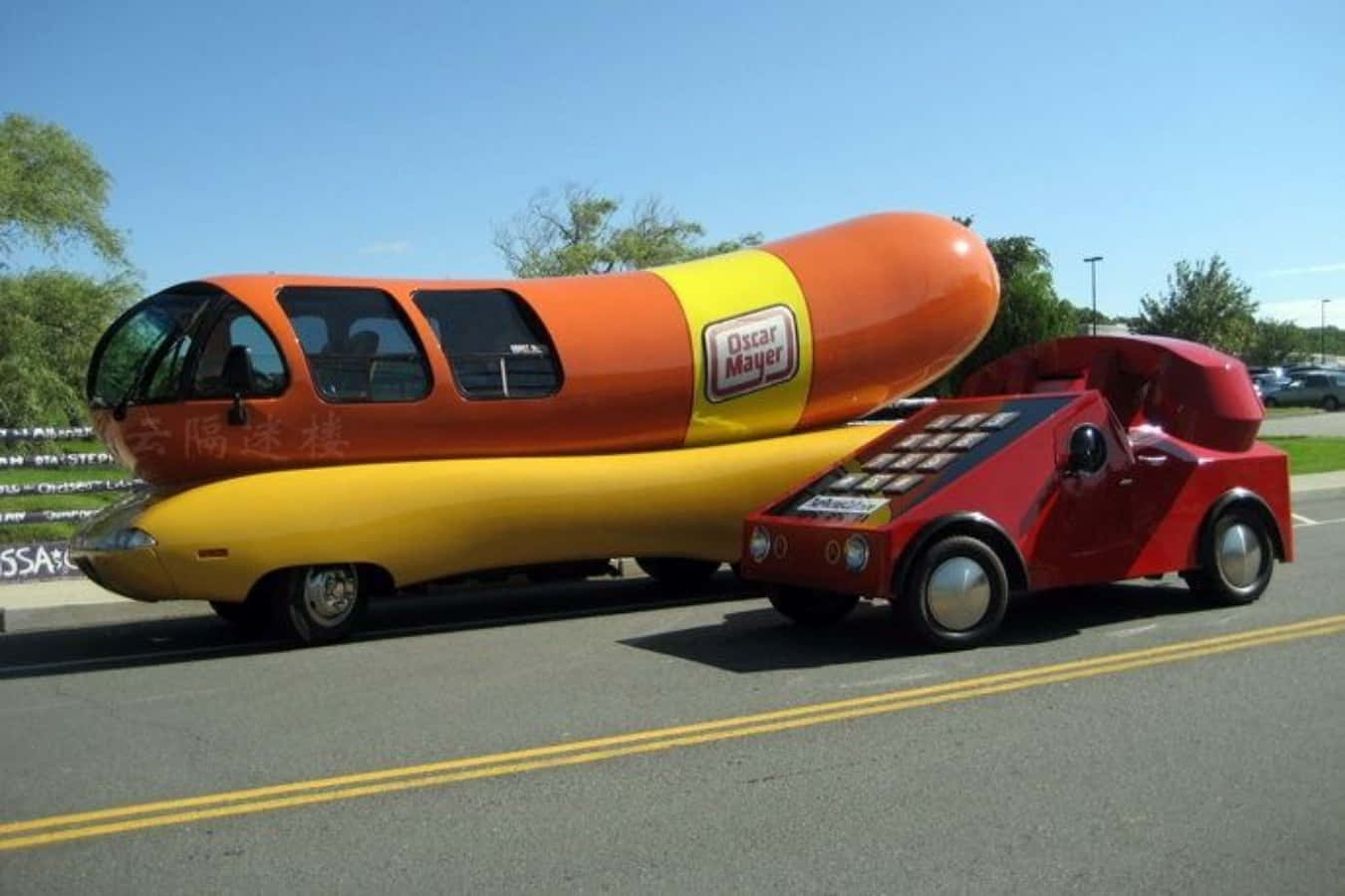 Funny Car 桌布 壁紙 — 一輛巨型奧斯卡·邁耶 Wienermobile 形狀像熱狗，帶紅色拖車，在陽光明媚的道路上行駛。