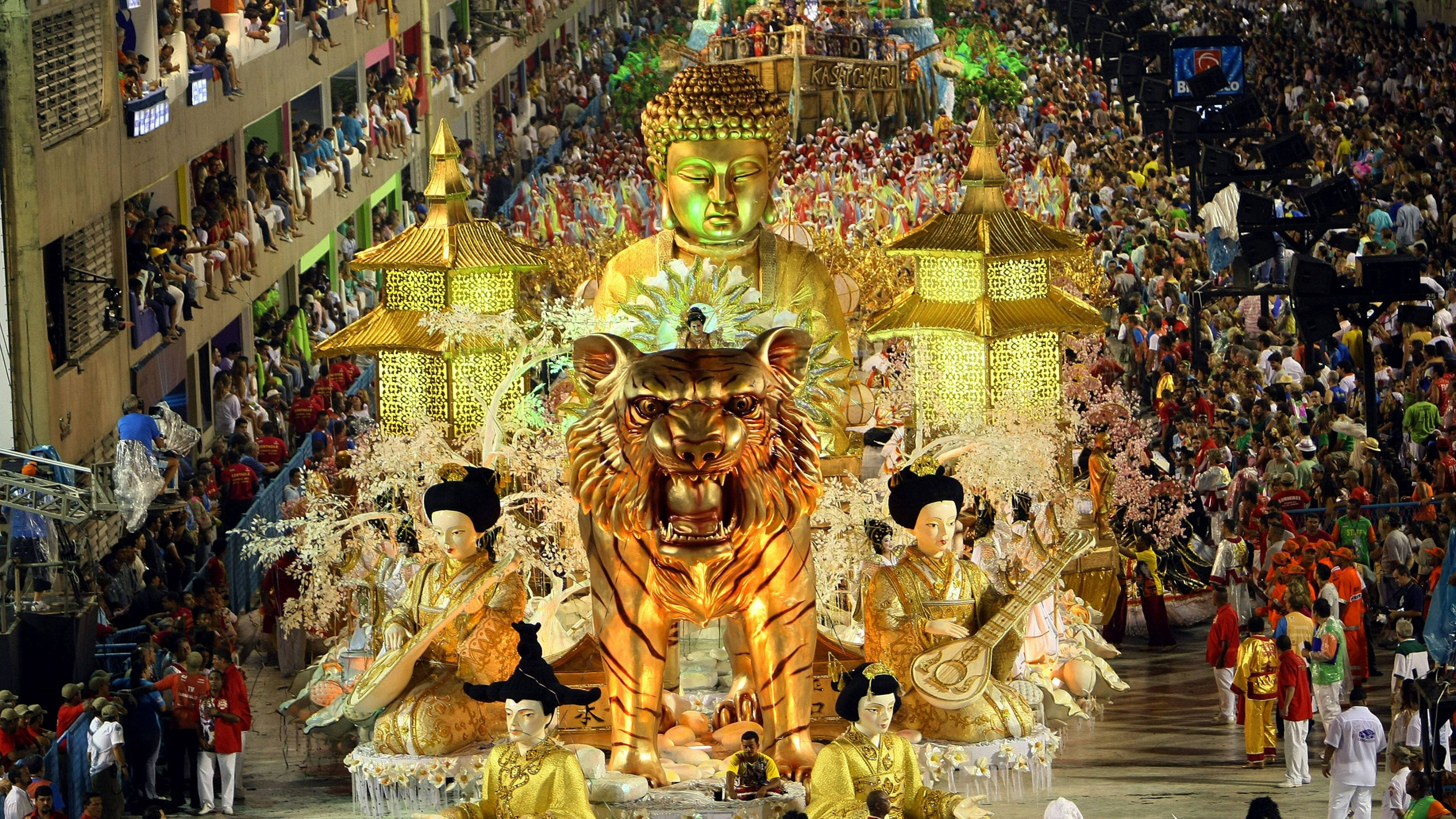 rio 桌布 壁紙 — 一輛巨大的金色虎頭花車，載有佛像和音樂家，在擁擠的夜間嘉年華街道上巡遊。