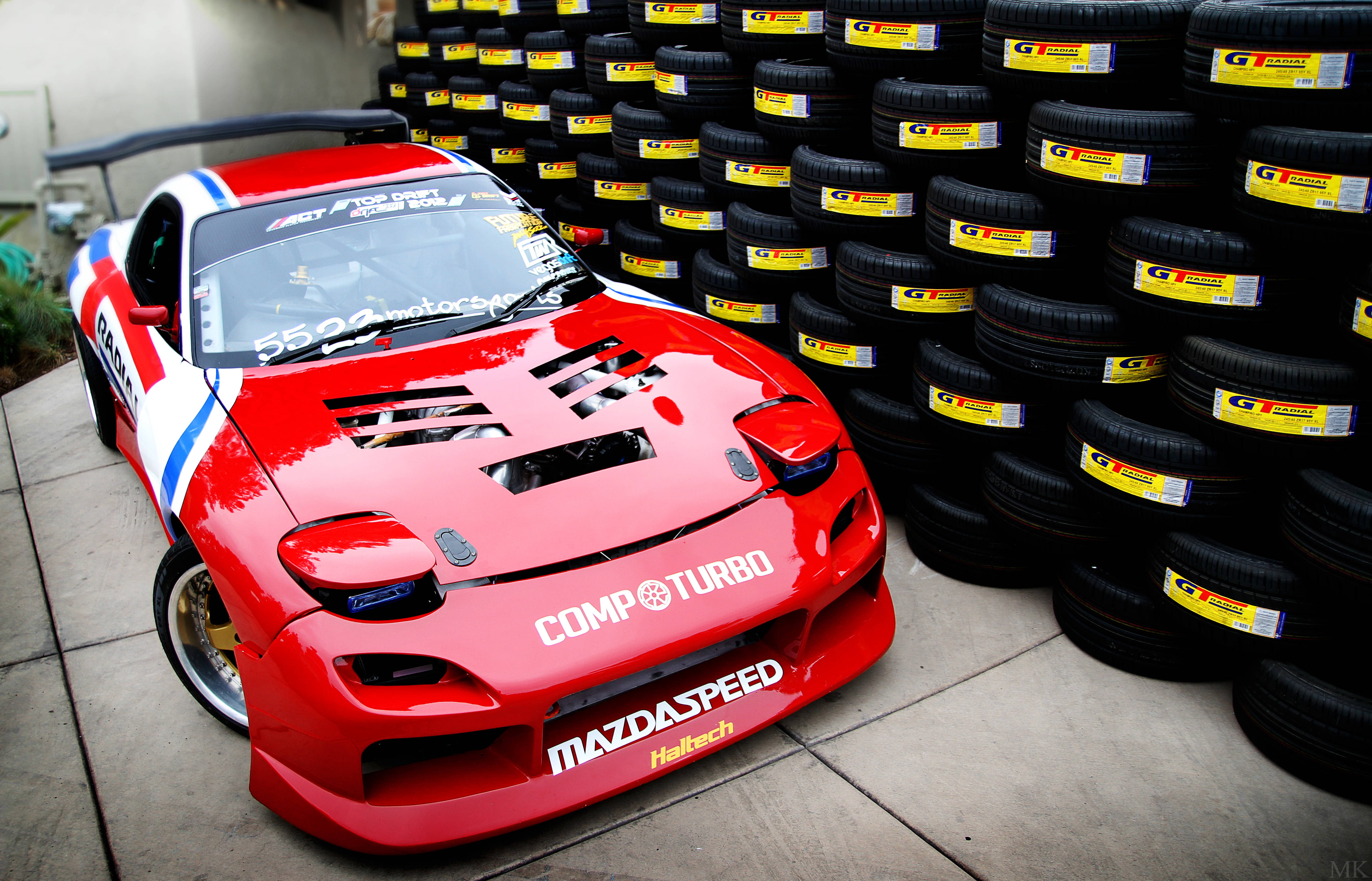 Mazda 桌布 壁紙 — 一輛帶有「CompTurbo」和「MazdaSpeed」貼紙的鮮紅色馬自達RX-7賽車，自豪地站在一堆黑色G Tire賽車輪胎旁邊。