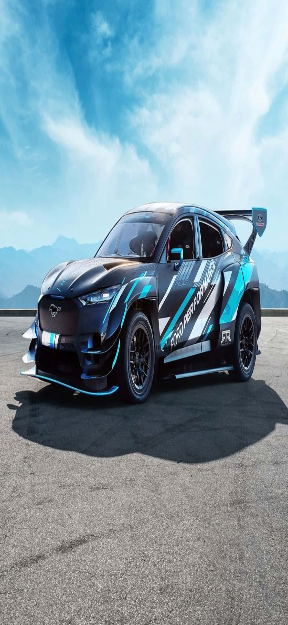 電玩遊戲 桌布 壁紙 — 一輛帶有 'Ford Performance' 貼紙的時尚黑色和藍綠色賽車停在鋪砌的道路上，背景是遠山和帶有薄雲的明亮藍天。