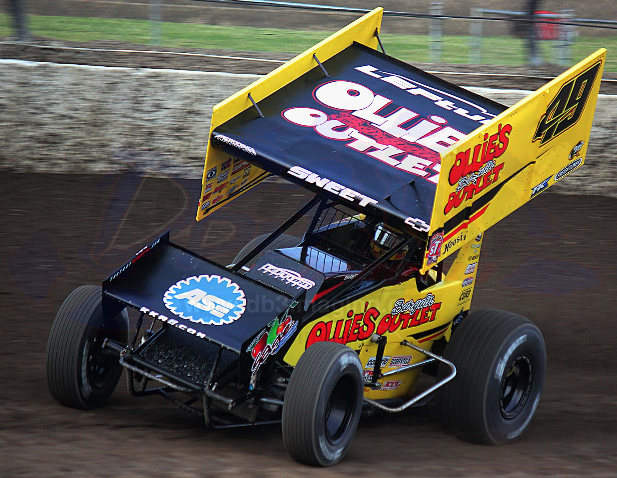 Sprint Car Racing 桌布 壁紙 — 一輛帶有「Ollie's Outlet」品牌的黃黑泥地賽車在泥濘的橢圓形賽道上飛馳。