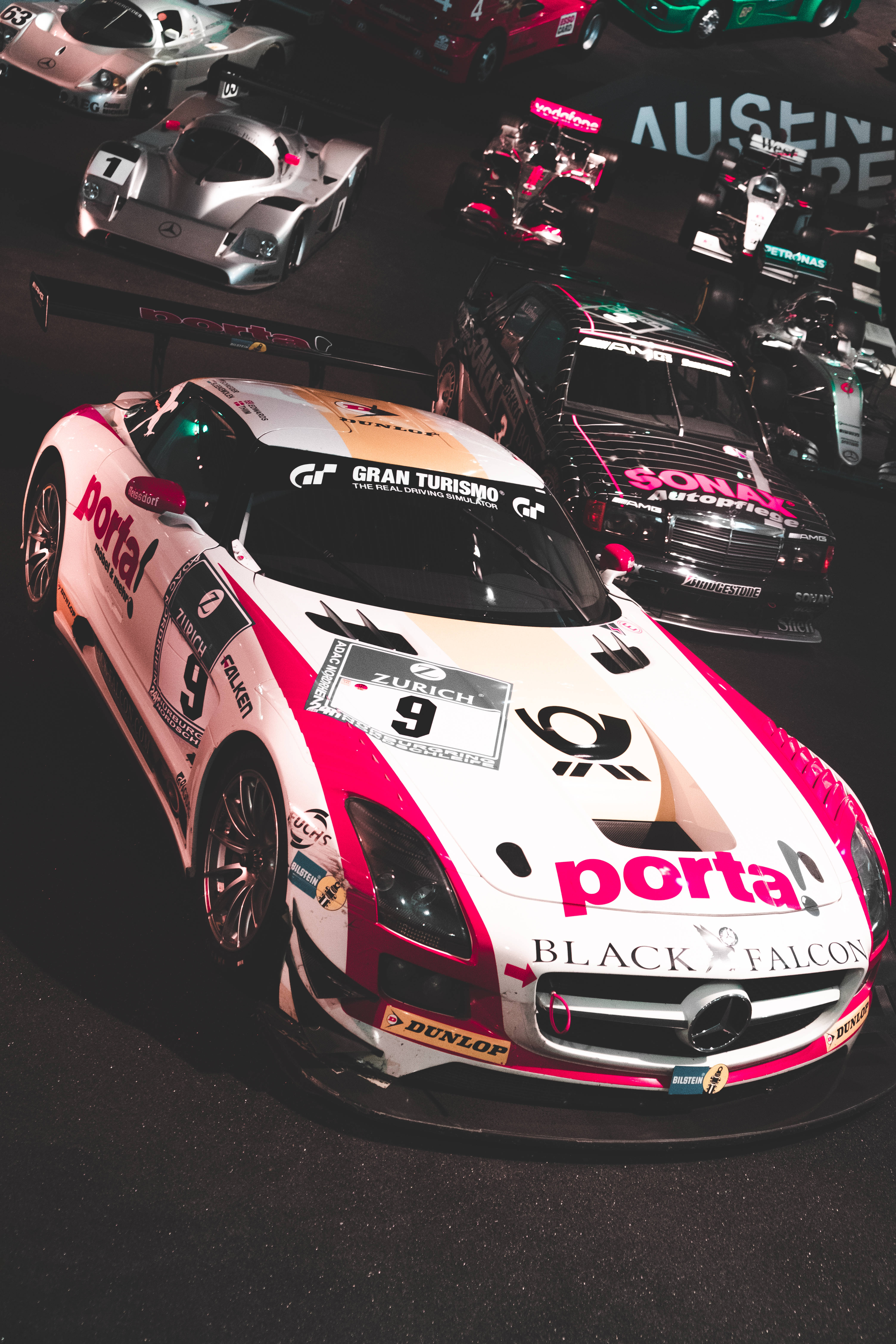 跑車 桌布 壁紙 — 一輛帶有「portal」品牌標誌的白色和粉紅色賽車，在一個黑暗的車庫展覽中，被其他高性能賽車包圍，佔據主導地位。