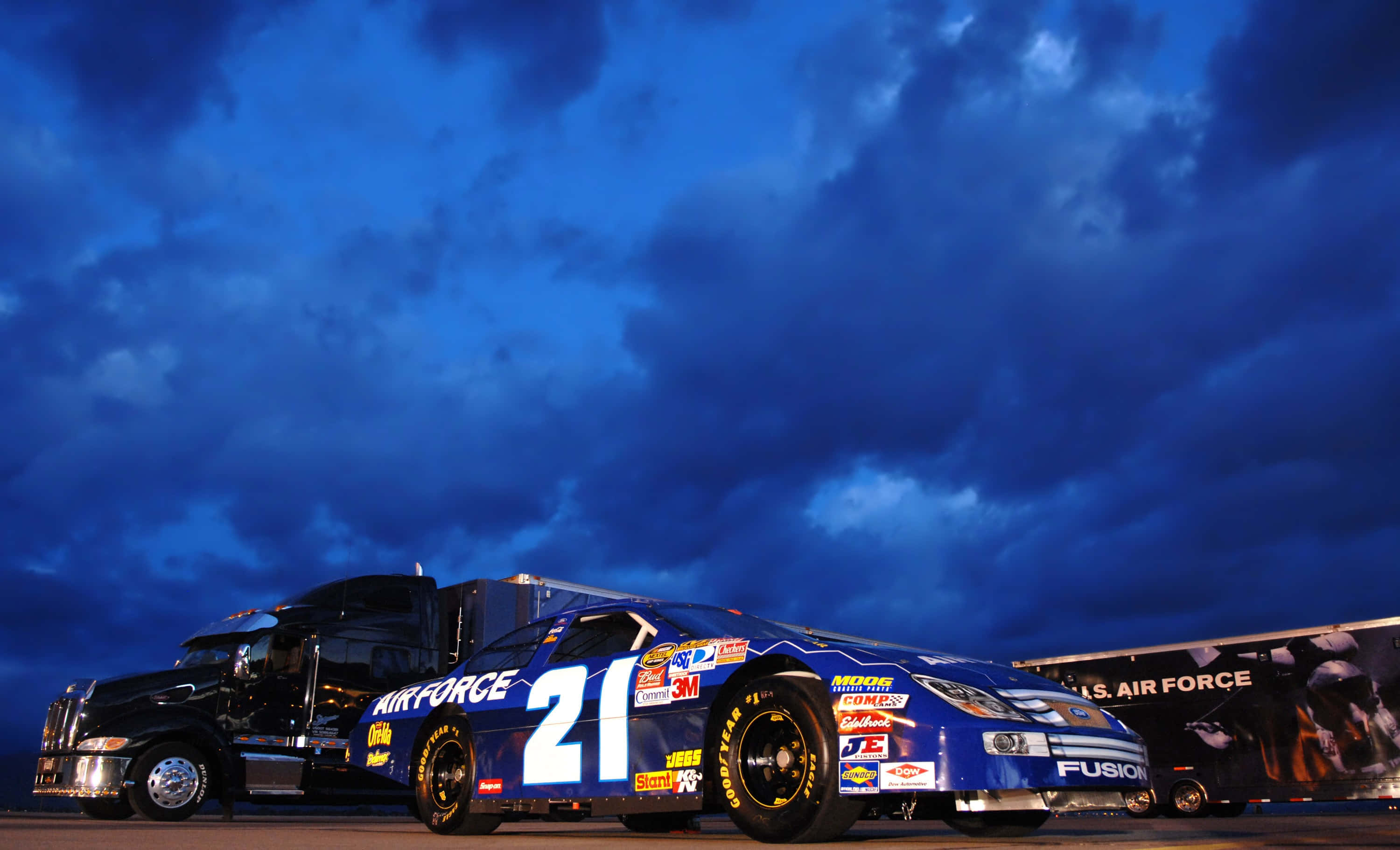 nascar 桌布 壁紙 — 一輛帶有21號和空軍品牌的藍色賽車停在一輛黑色半掛卡車旁邊，背景是戲劇性的暮色天空。