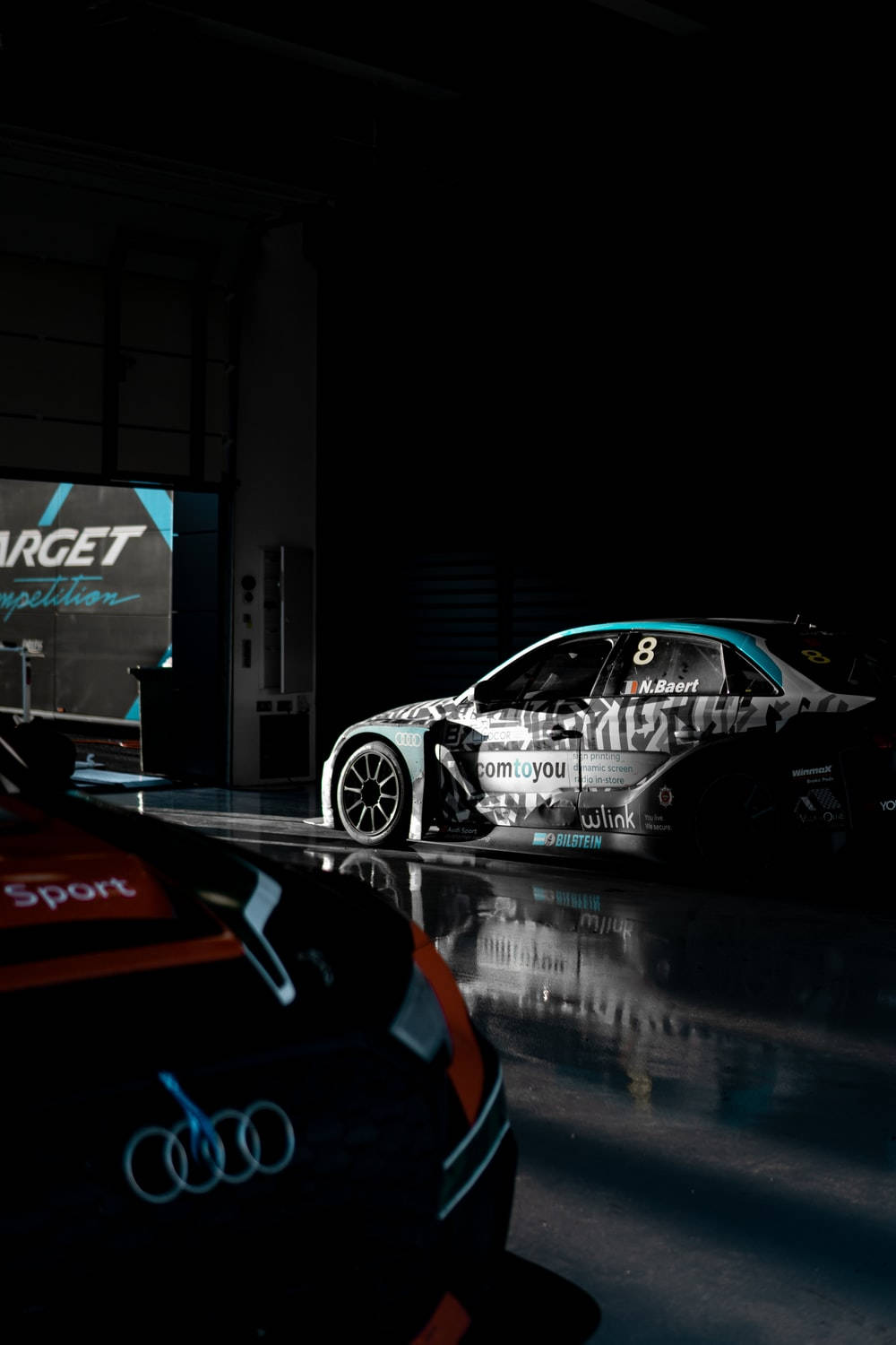 Auto Racing 桌布 壁紙 — 一輛帶有8號和贊助商貼紙的賽車在黑暗的車庫中戲劇性地被照亮，地面濕滑反光。