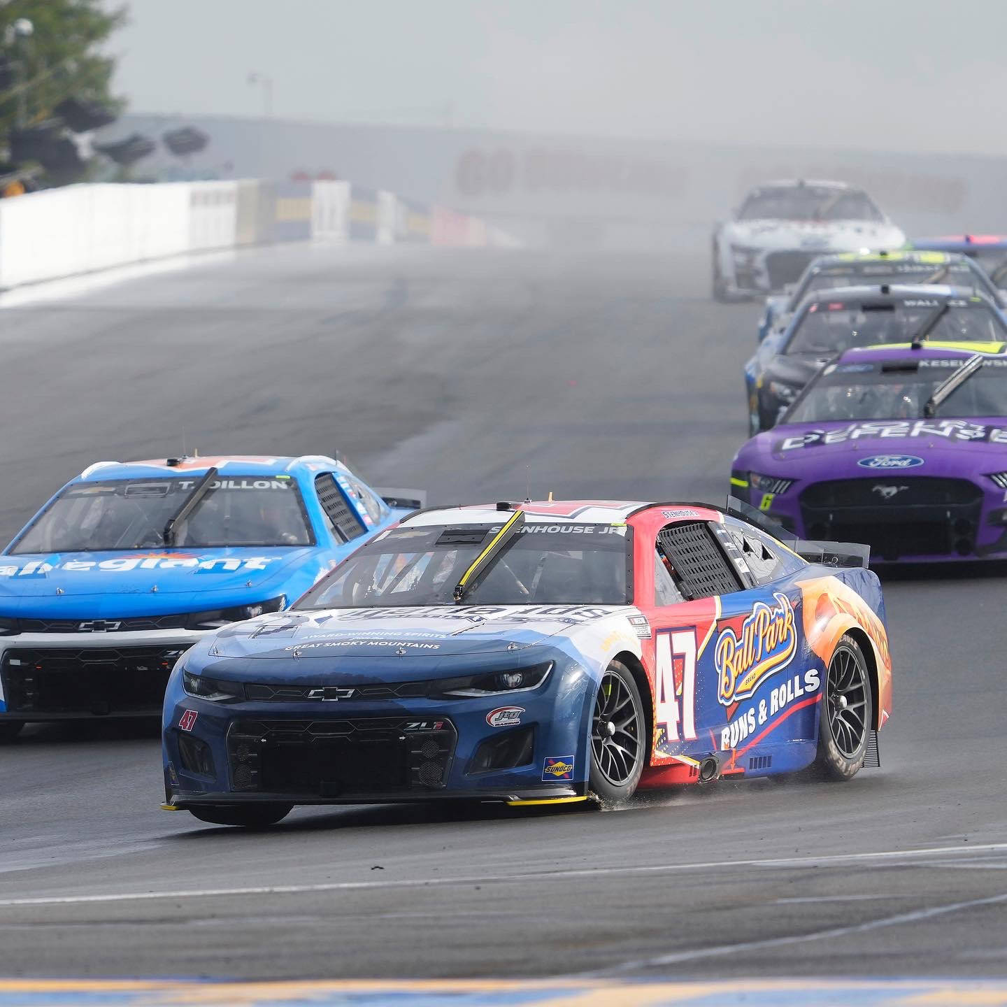 Racing 桌布 壁紙 — 一輛帶有Ball Park標誌的藍色NASCAR賽車在潮濕的賽道上飛馳，競爭對手在霧氣瀰漫的比賽中落後。