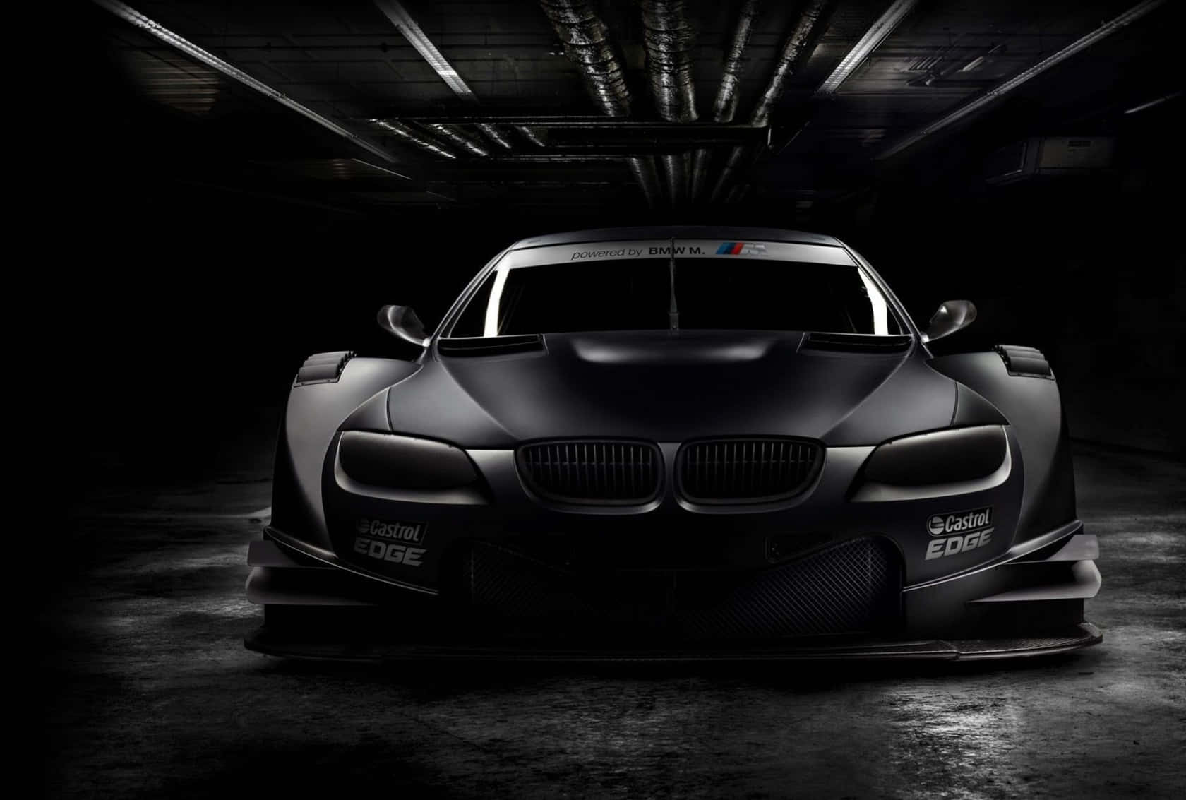 Bmw M Series 桌布 壁紙 — 一輛帶有Castrol EDGE品牌的消光黑色BMW賽車在黑暗的工業車庫中被戲劇性地照亮。